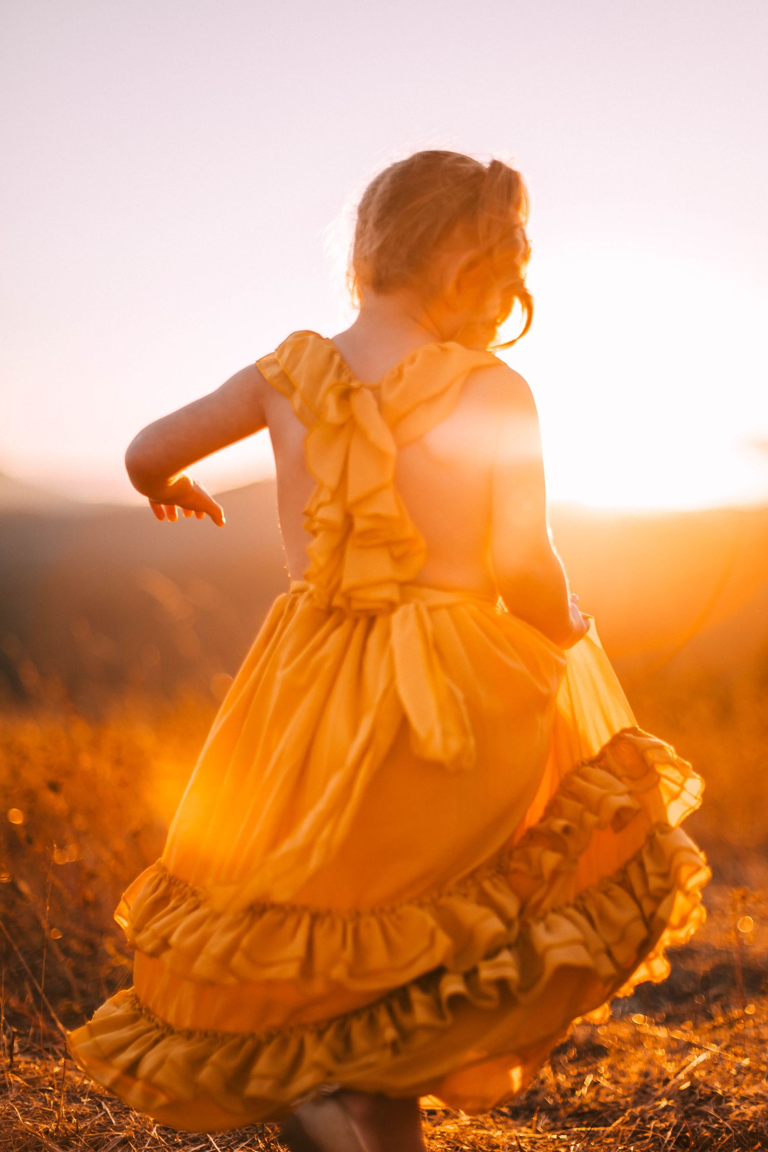 A young child in a yellow dress twirls in a field at sunset, creating a dreamy silhouette against the golden light.