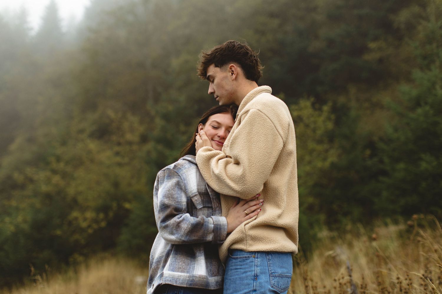 A couple embraces tenderly in a misty autumn field surrounded by trees and tall grass.