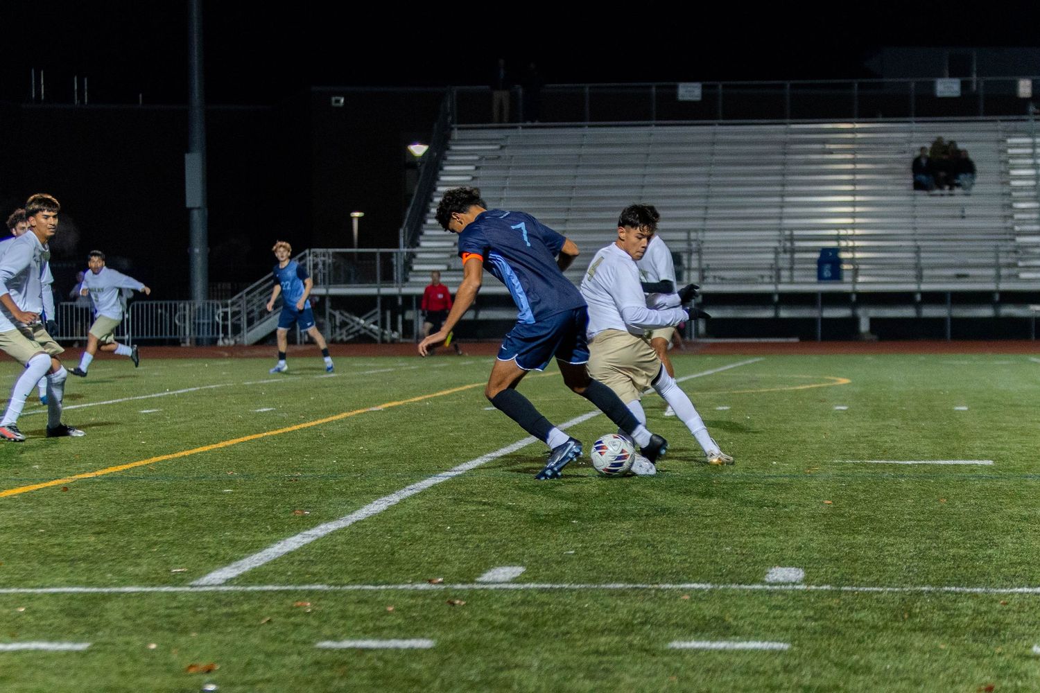 Soccer players compete for the ball during an intense match on an outdoor turf field under stadium lights.