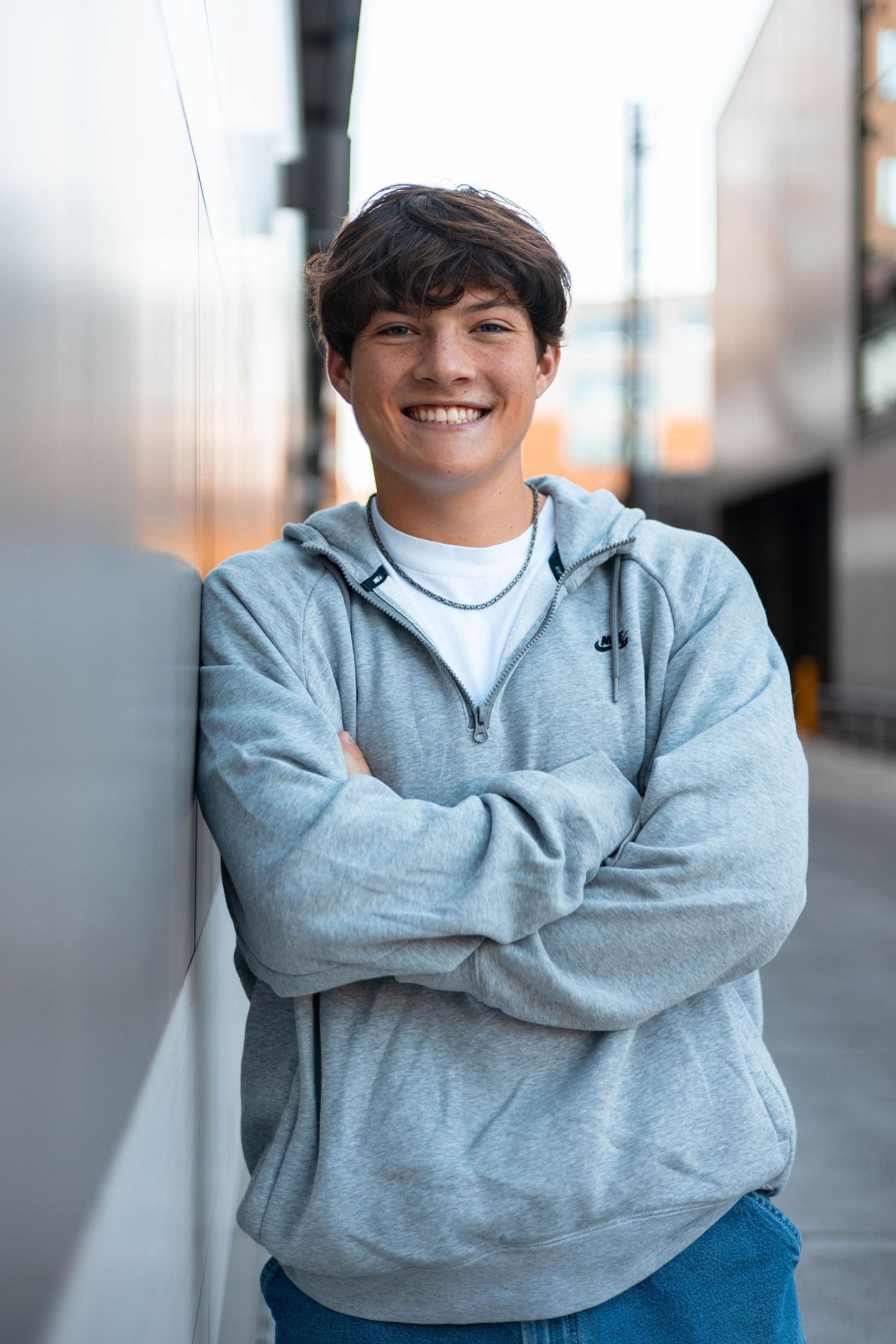 Young person in a gray hoodie and jeans smiles warmly while leaning against a wall outdoors.