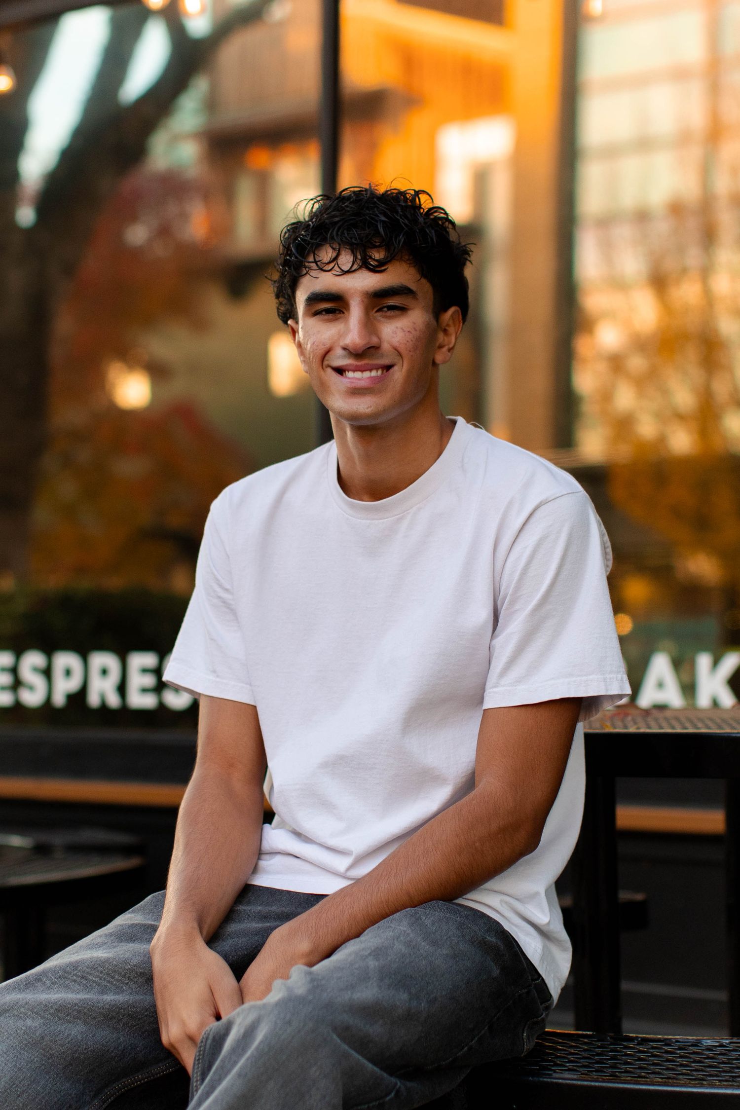 A person wearing a white t-shirt and gray pants sits outdoors on a bench with warm lighting and storefronts in background.