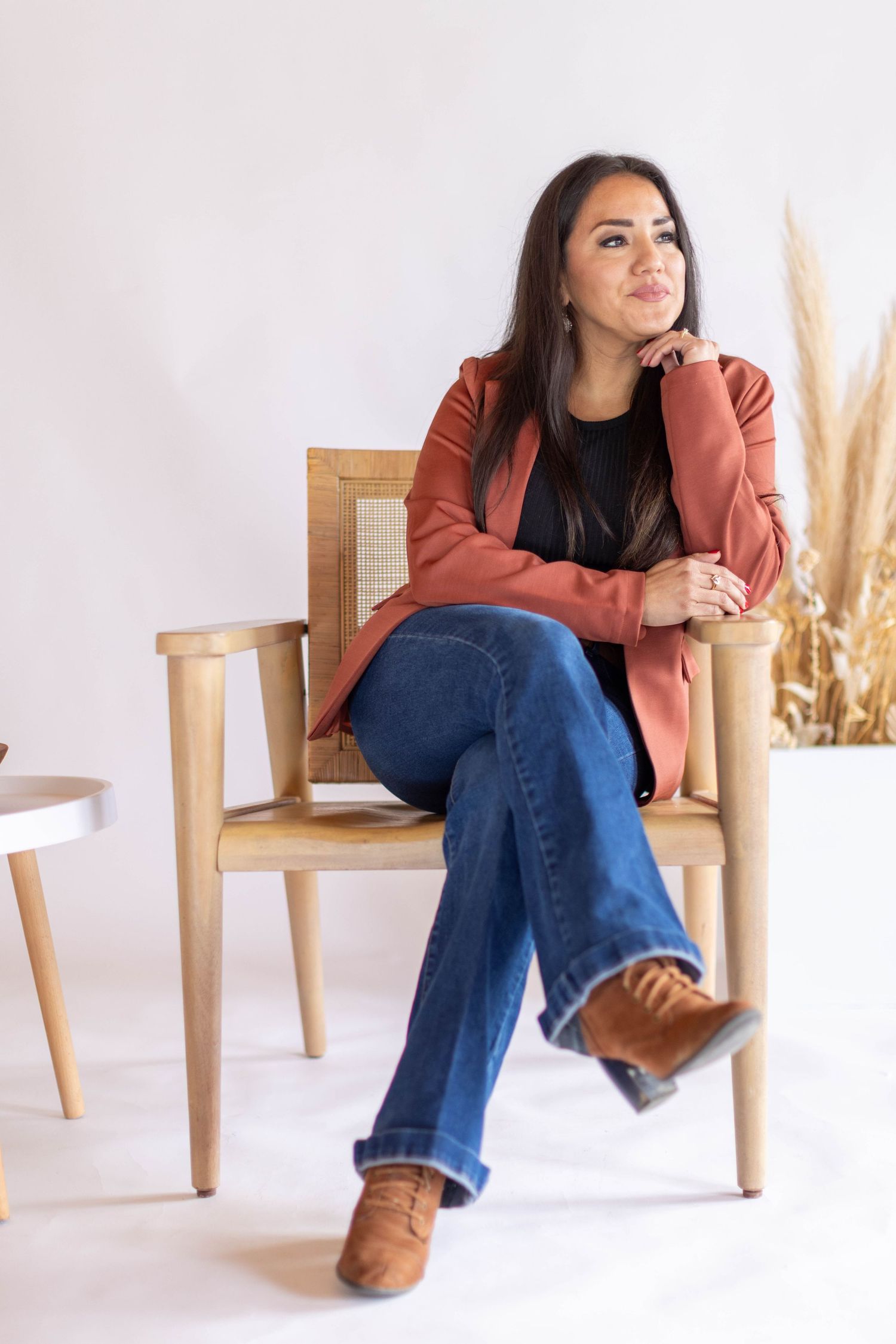 A woman in a rust-colored sweater and blue jeans sits casually on a wooden chair against a white backdrop.