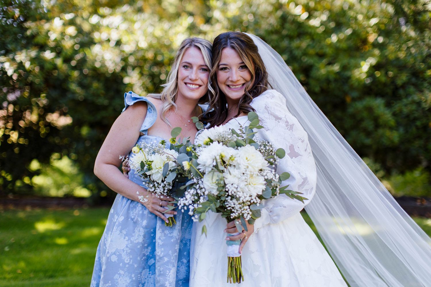 Bridesmaids in light blue dresses gather with bride in white gown holding white floral bouquets in sunny garden setting.