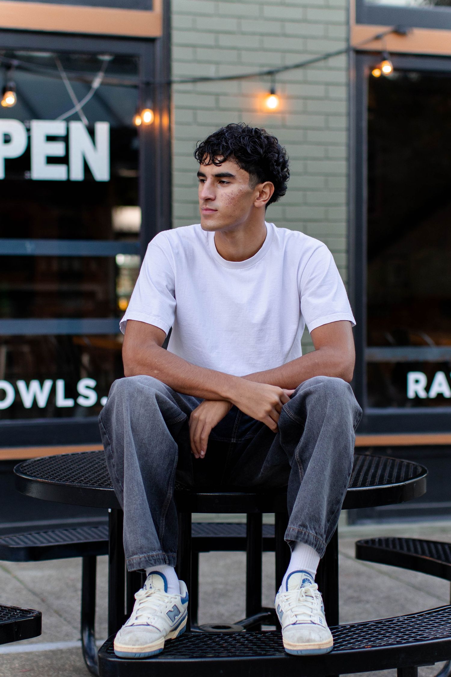 Person in white t-shirt and jeans sits on black metal bench outside storefront with illuminated lights.
