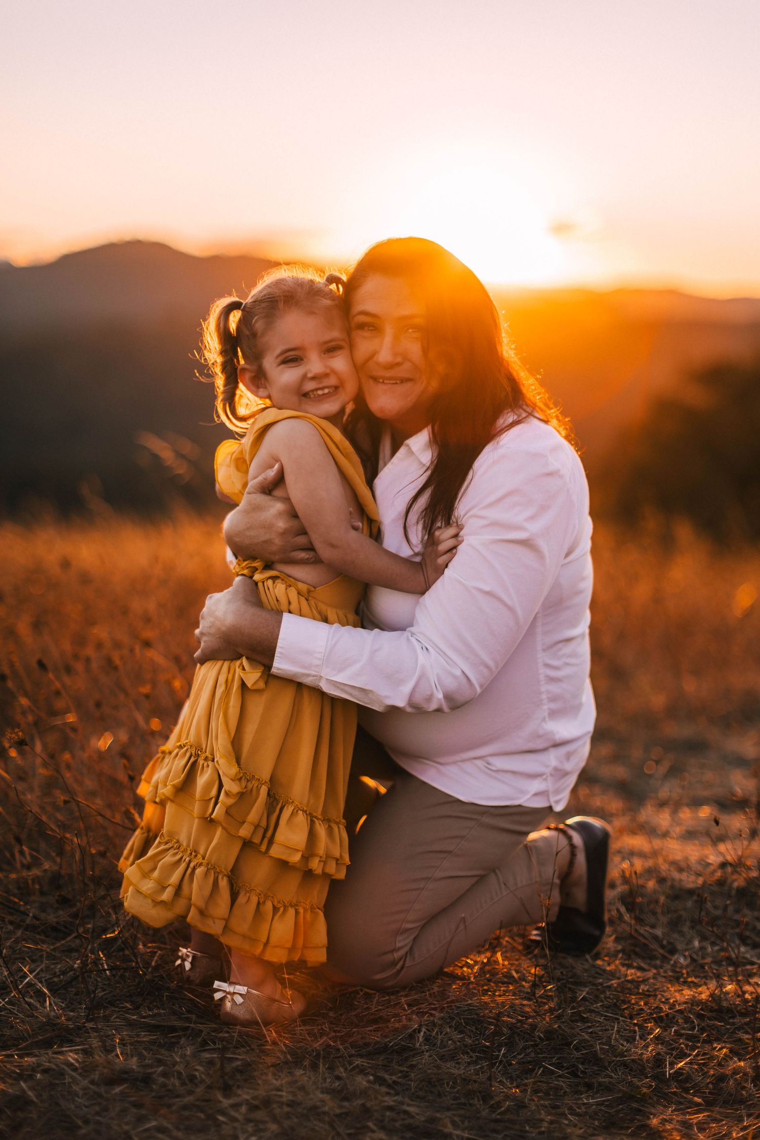 Two figures share a tender embrace in a field during golden hour, backlit by the warm glow of sunset.