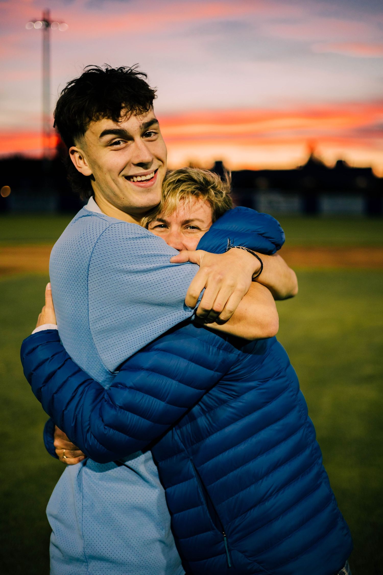 Two people share a warm embrace outdoors during a vibrant sunset with orange and pink skies in the background.