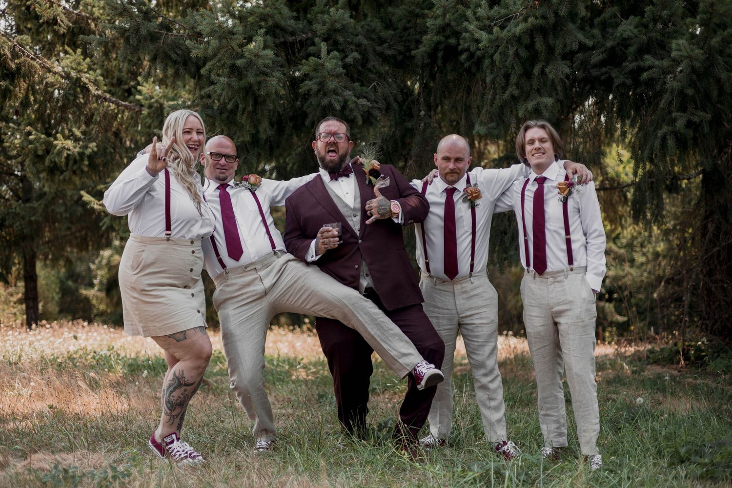 Wedding party members in white and burgundy attire pose playfully together in an outdoor garden setting.