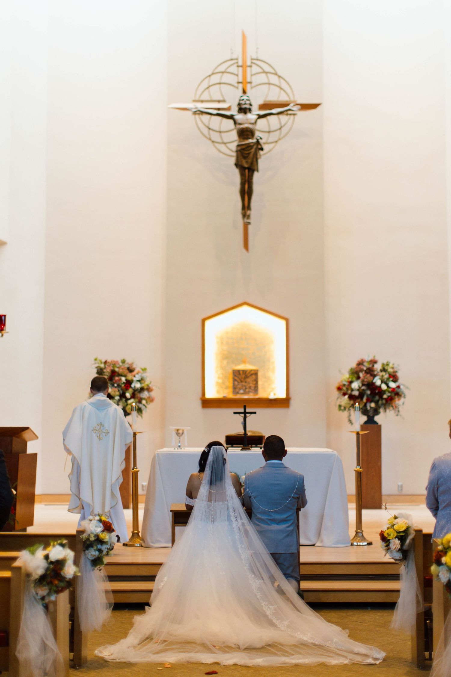 A wedding ceremony in a modern church with white walls, floral arrangements, and a hanging crucifix.