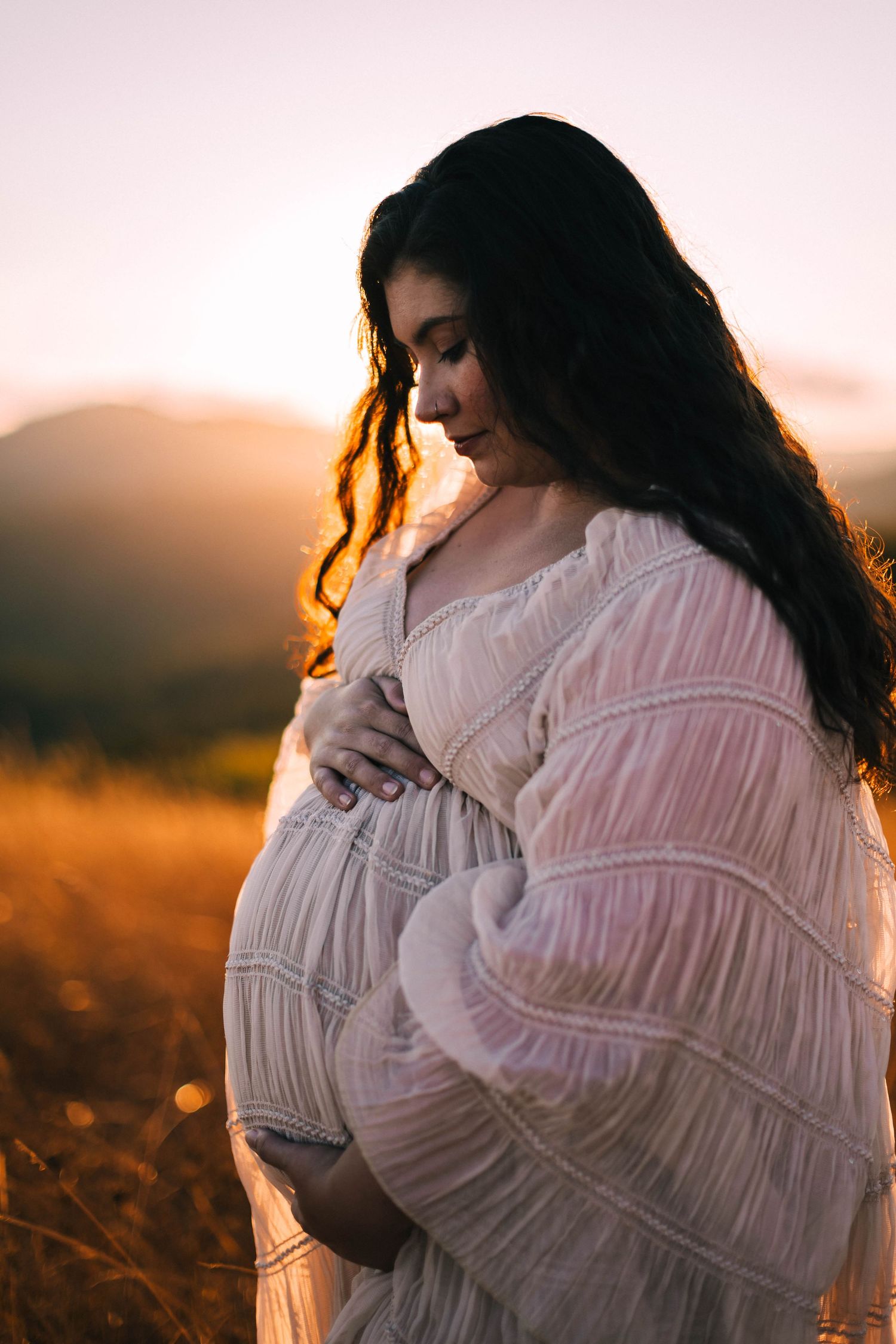 Silhouette of a pregnant woman in a white dress standing in golden sunlight during sunset.