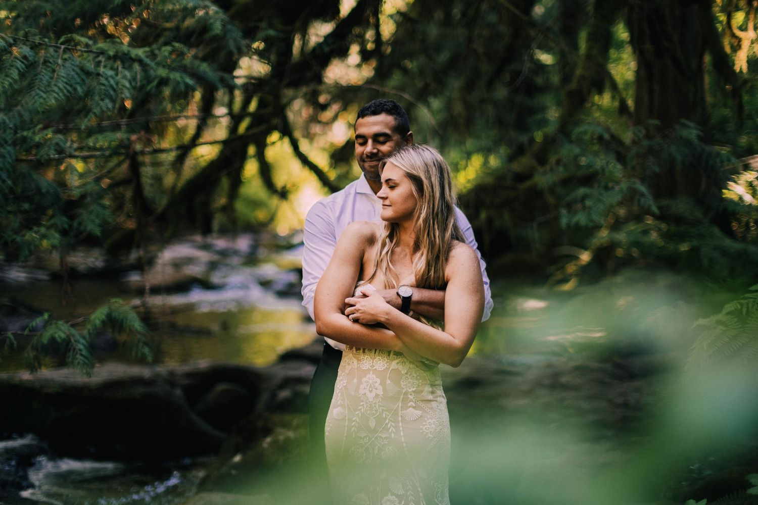 Romantic couple embraces near a tranquil forest stream during golden hour photography session.
