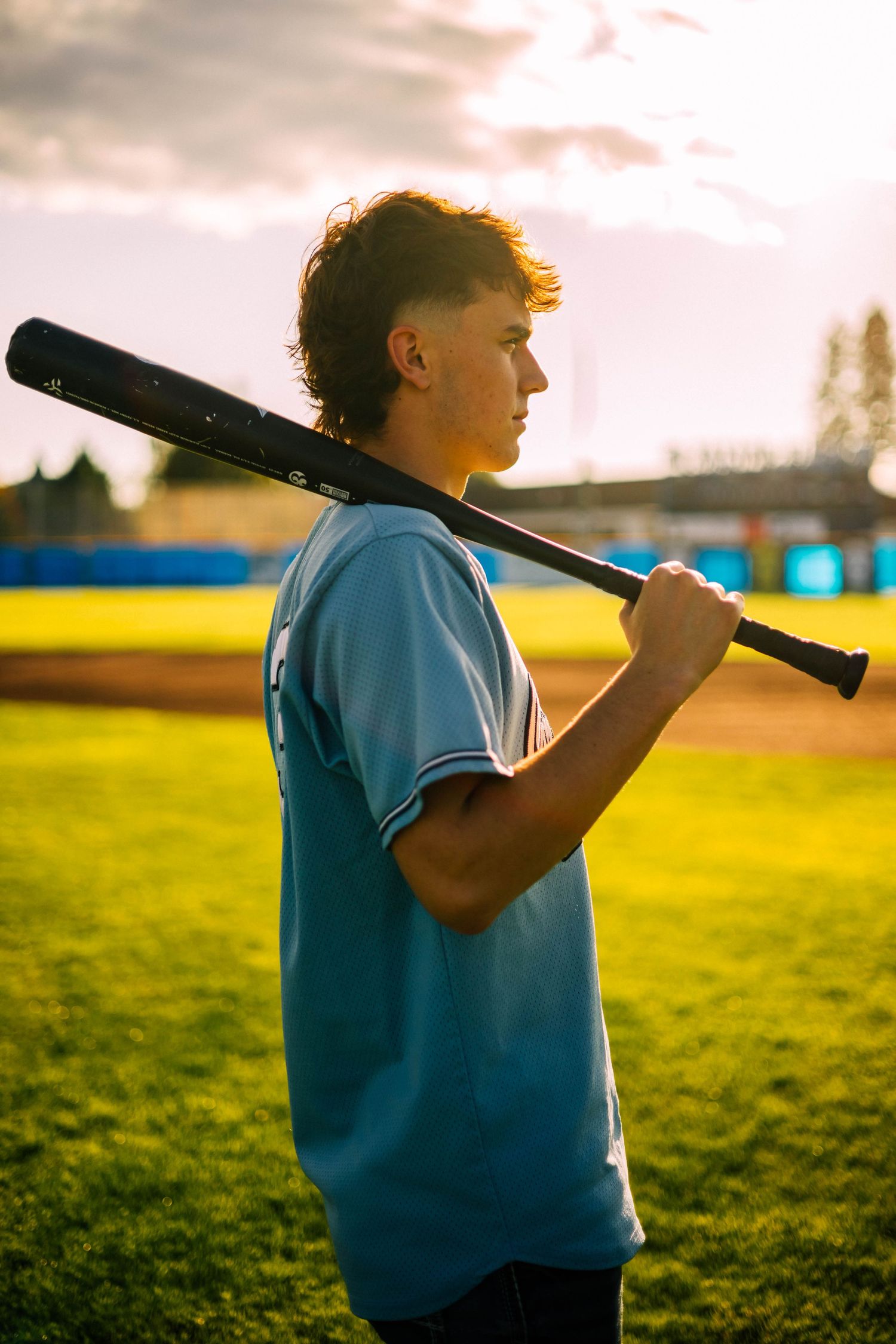 Baseball player in blue jersey holds bat over shoulder at sunset on grassy field.