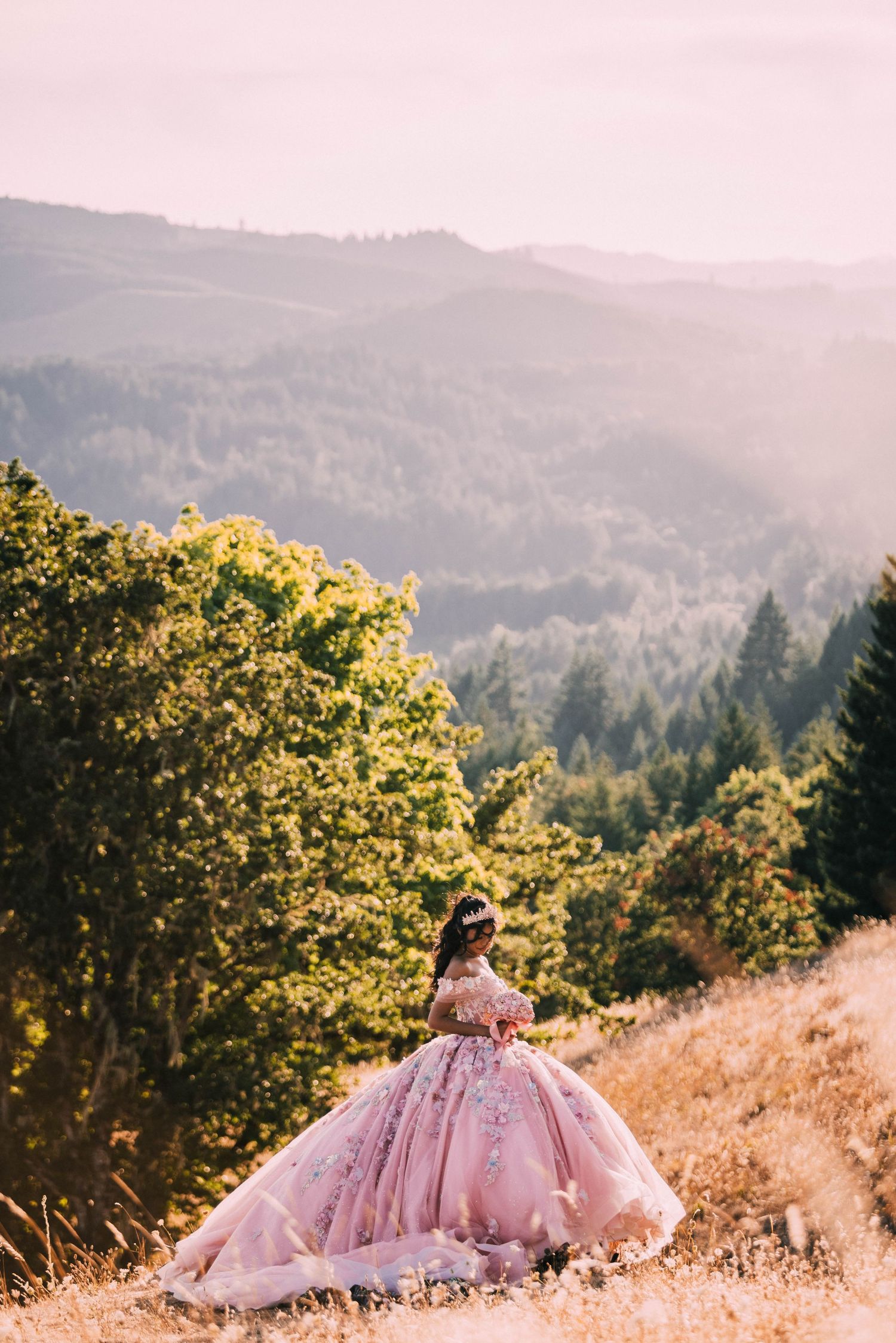 A person in a flowing pink ball gown twirls on a hilltop against a backdrop of mountains and forests at sunset.
