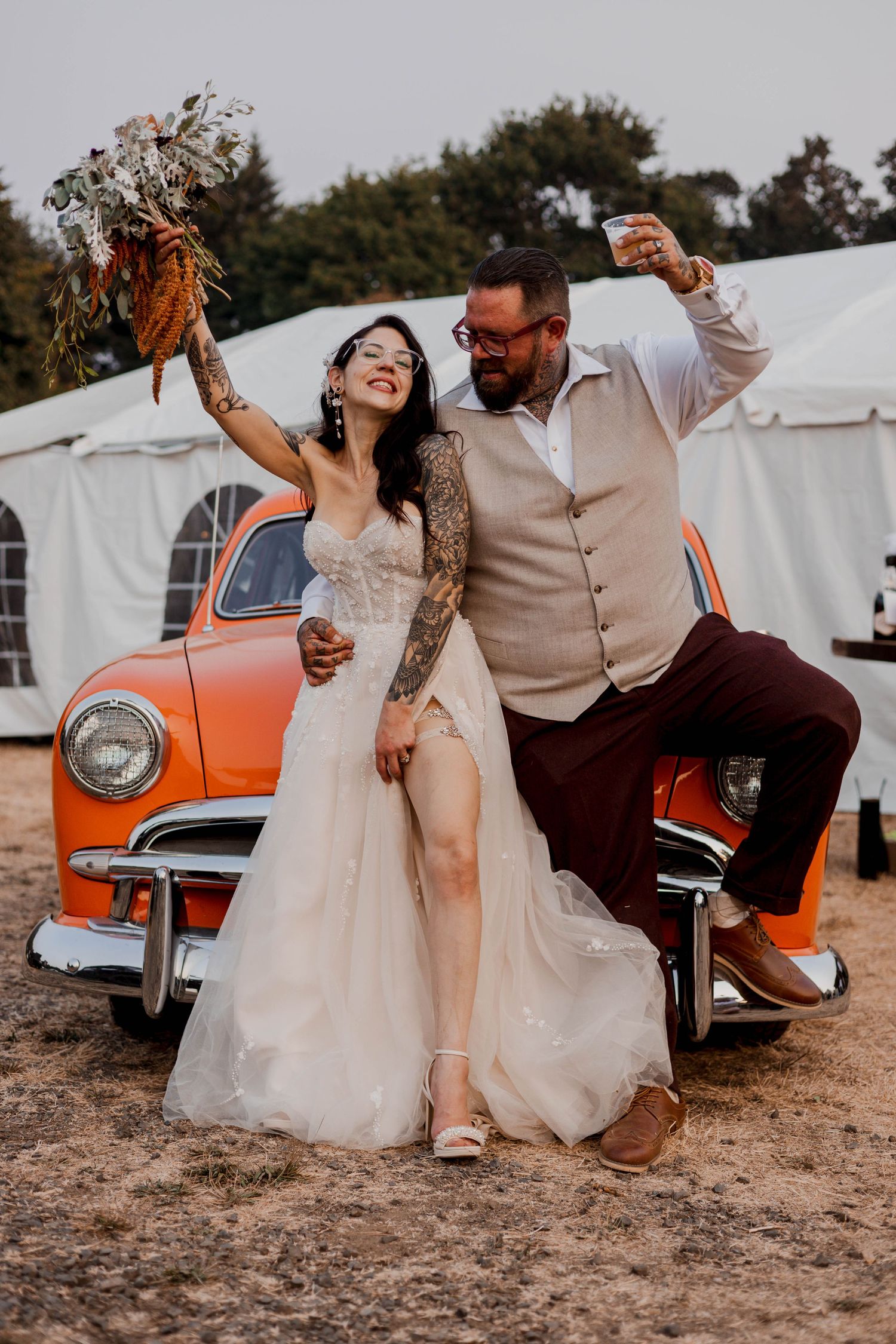 Playful wedding couple poses and dances near a classic orange vintage car in casual outdoor portraits.