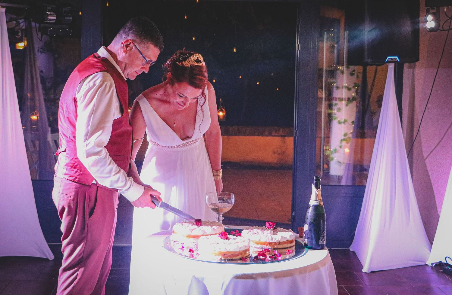 Les mariés découpant ensemble leur gâteau de mariage à trois étages, décoré de pétales de roses rouges, lors de la réception nocturne