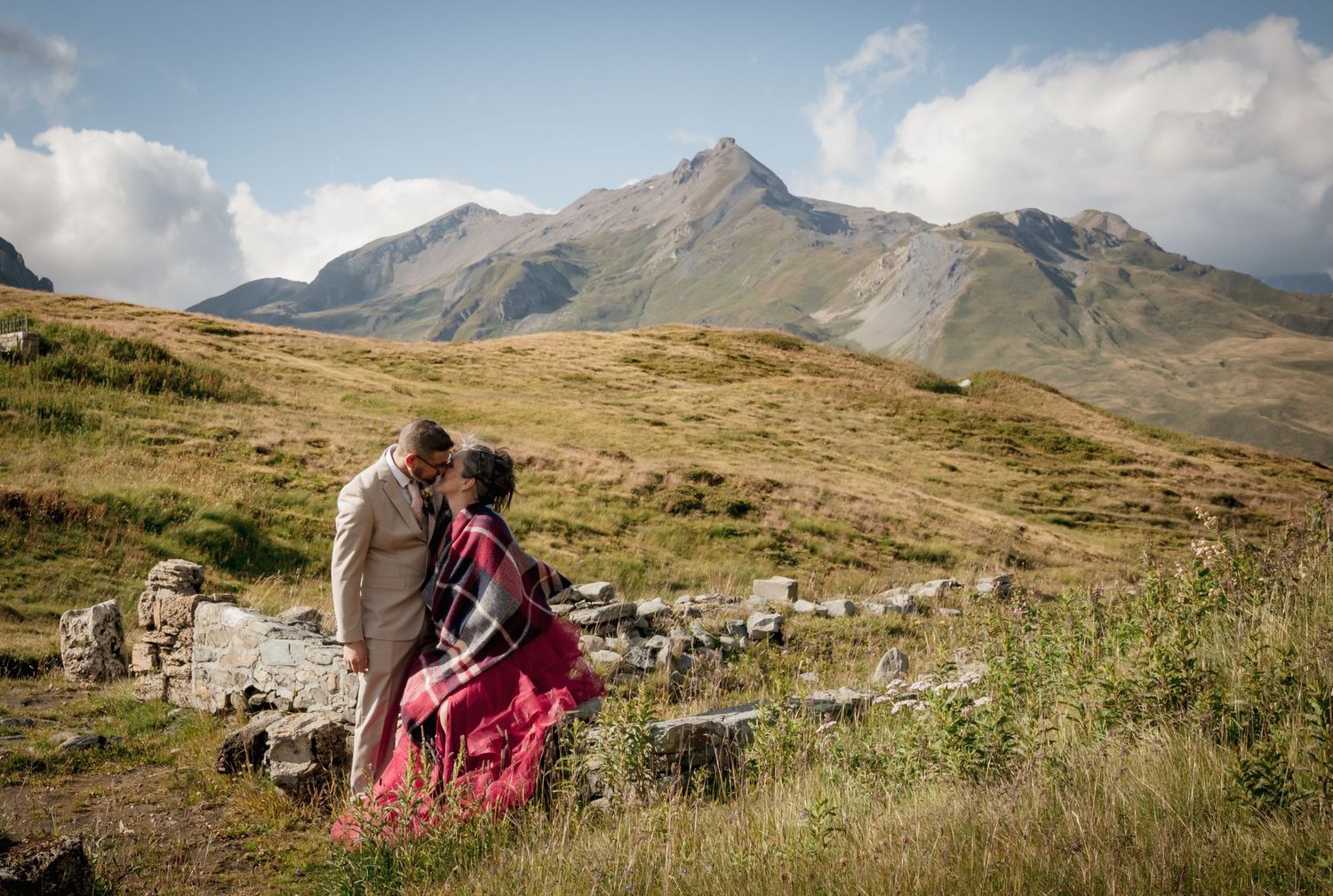 Couple marié s'embrassant en montagne devant un sommet rocheux, mariée en robe rouge avec plaid écossais, décor naturel alpin.