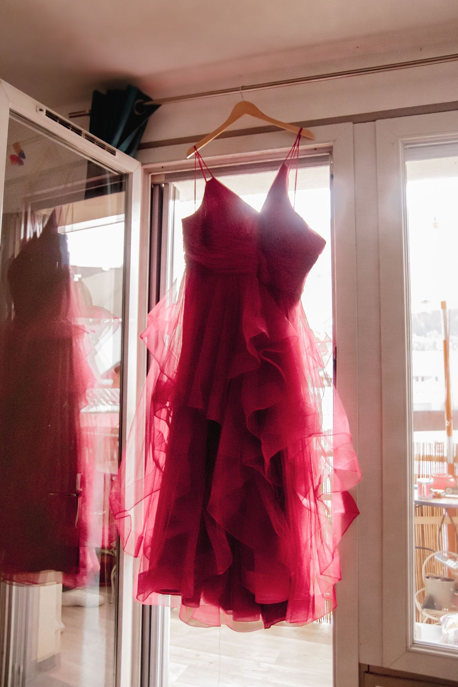 Robe de mariée rouge intense à volants en tulle, suspendue sur un cintre devant une fenêtre lumineuse.