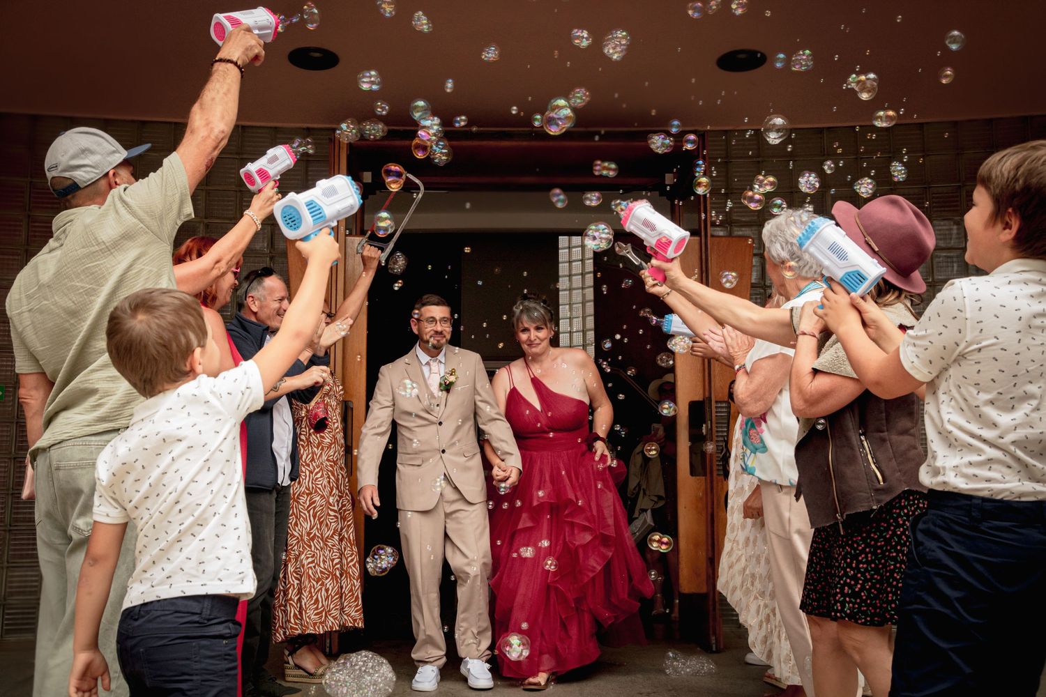 Sortie de mariage festive : couple sous une pluie de bulles de savon lancées par les invités, mariée en robe rouge éclatante et souriante