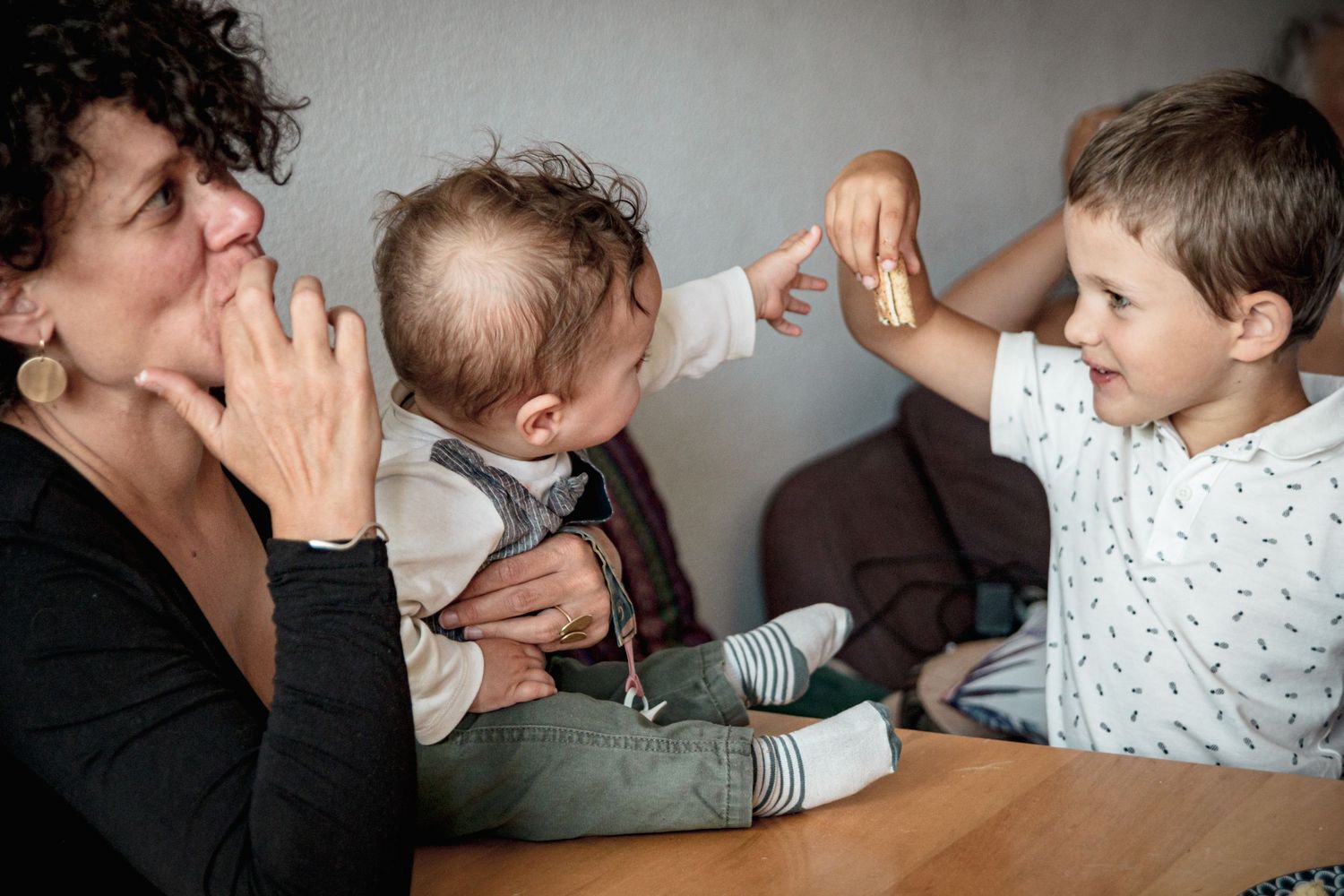 Moment familial tendre entre une maman, son bébé et un enfant, partageant un biscuit autour d'une table, ambiance chaleureuse et naturelle