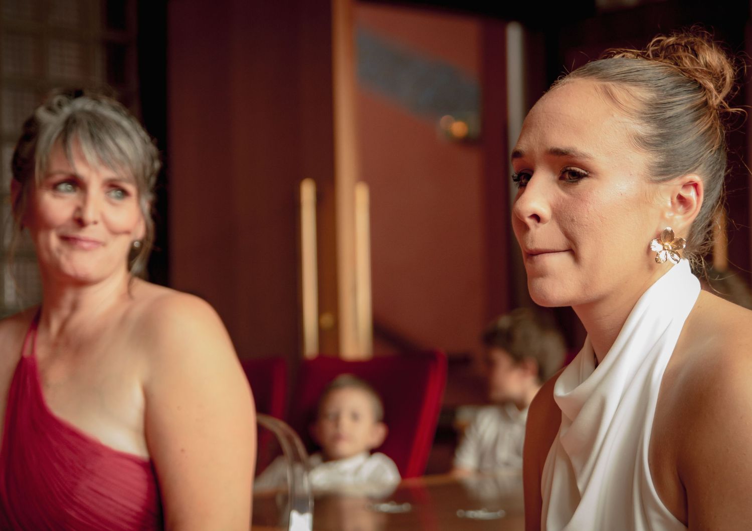 Portrait d'une invitée de mariage élégante avec boucles d'oreilles fleuries, la mariée en robe rouge en arrière-plan lors de la réception