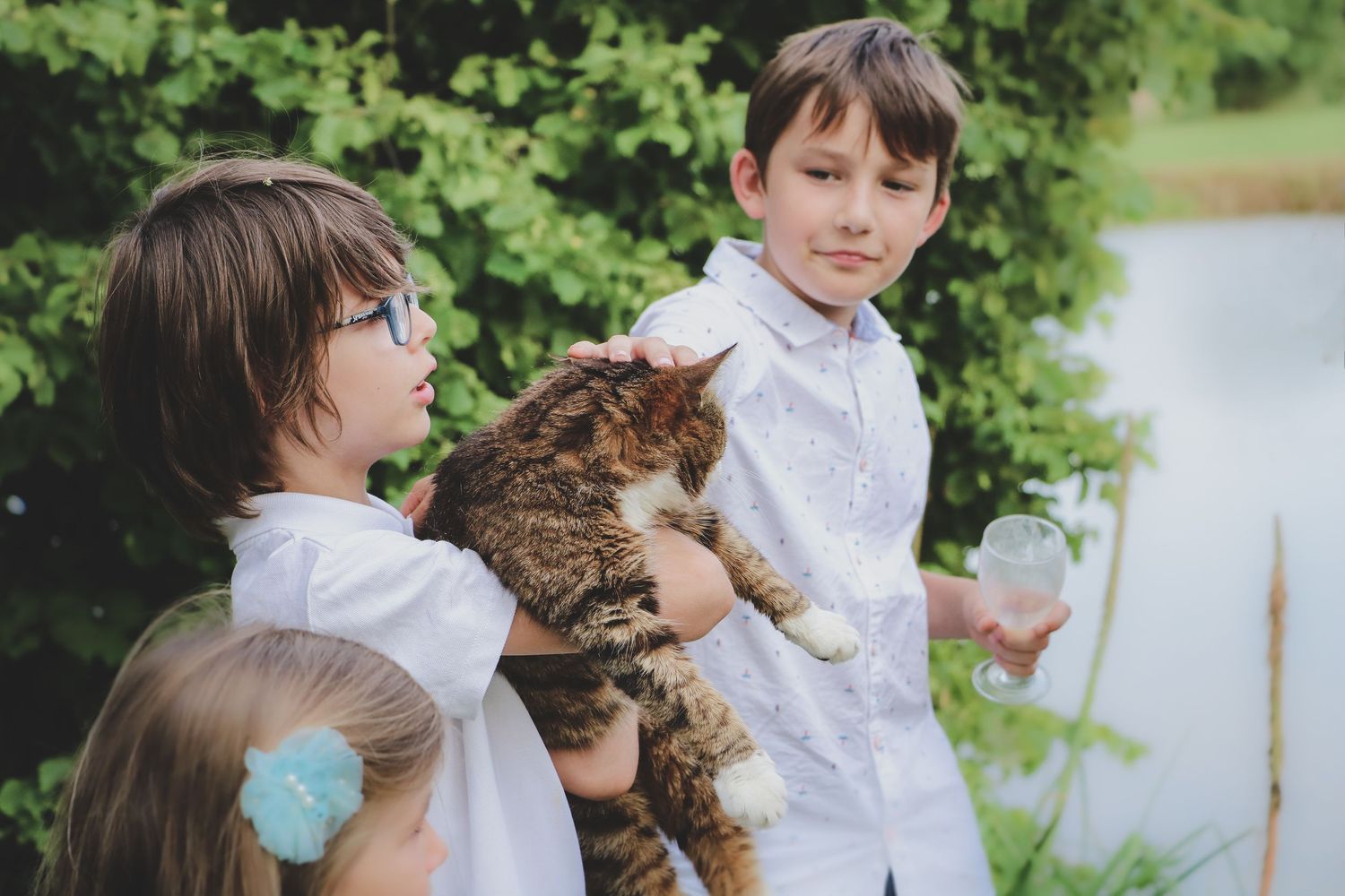 Enfants en tenue de cérémonie caressant un chat lors d’un mariage en extérieur, ambiance familiale et naturelle dans un jardin verdoyant