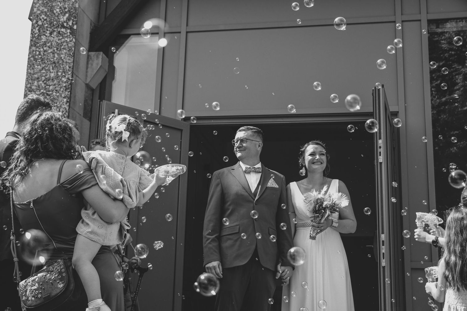 Couple de mariés souriant sous une pluie de bulles de savon à la sortie de la mairie, ambiance joyeuse et festive entourée des invités