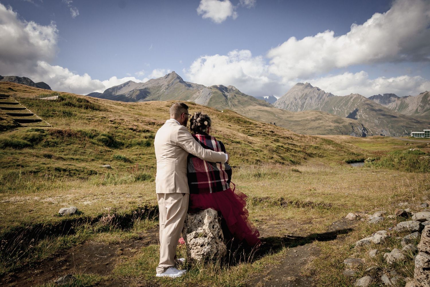 Couple de mariés de dos face aux sommets des Alpes, mariée en robe rouge et plaid tartan, paysage de montagne sous un ciel bleu et nuageux.
