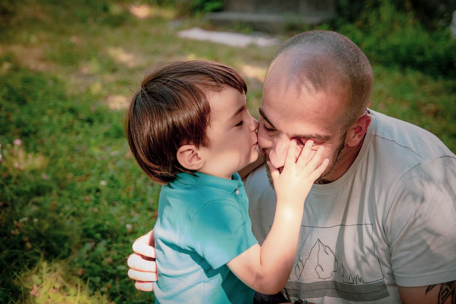 Un petit garçon embrasse son papa sur la joue en lui touchant le visage avec sa petite main, au coeur de leur jardin, photographe de famille