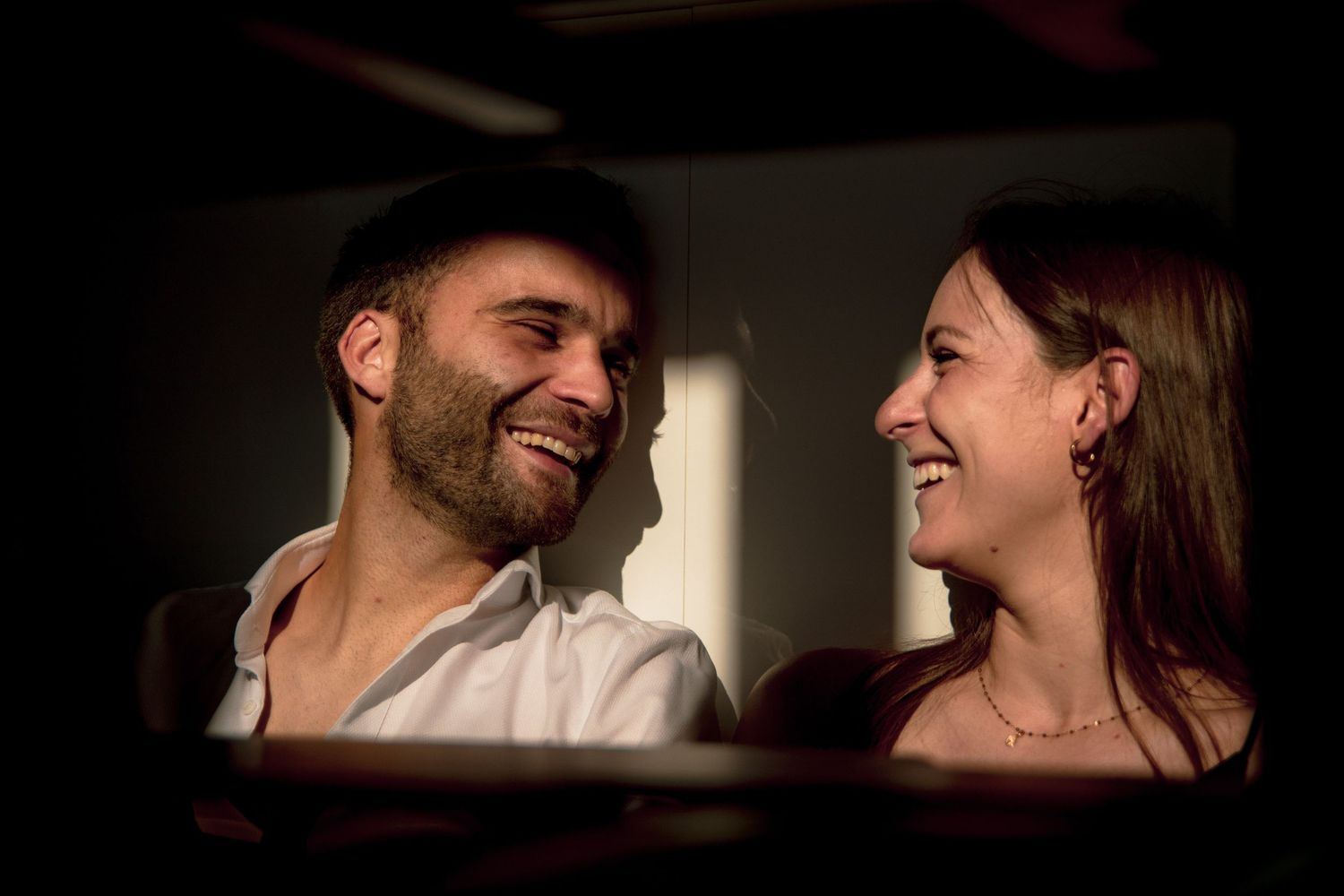 Couple rire aux éclats et complice lors d'une séance photo en intérieur, ambiance chaleureuse et naturelle pour une séance d'engagement