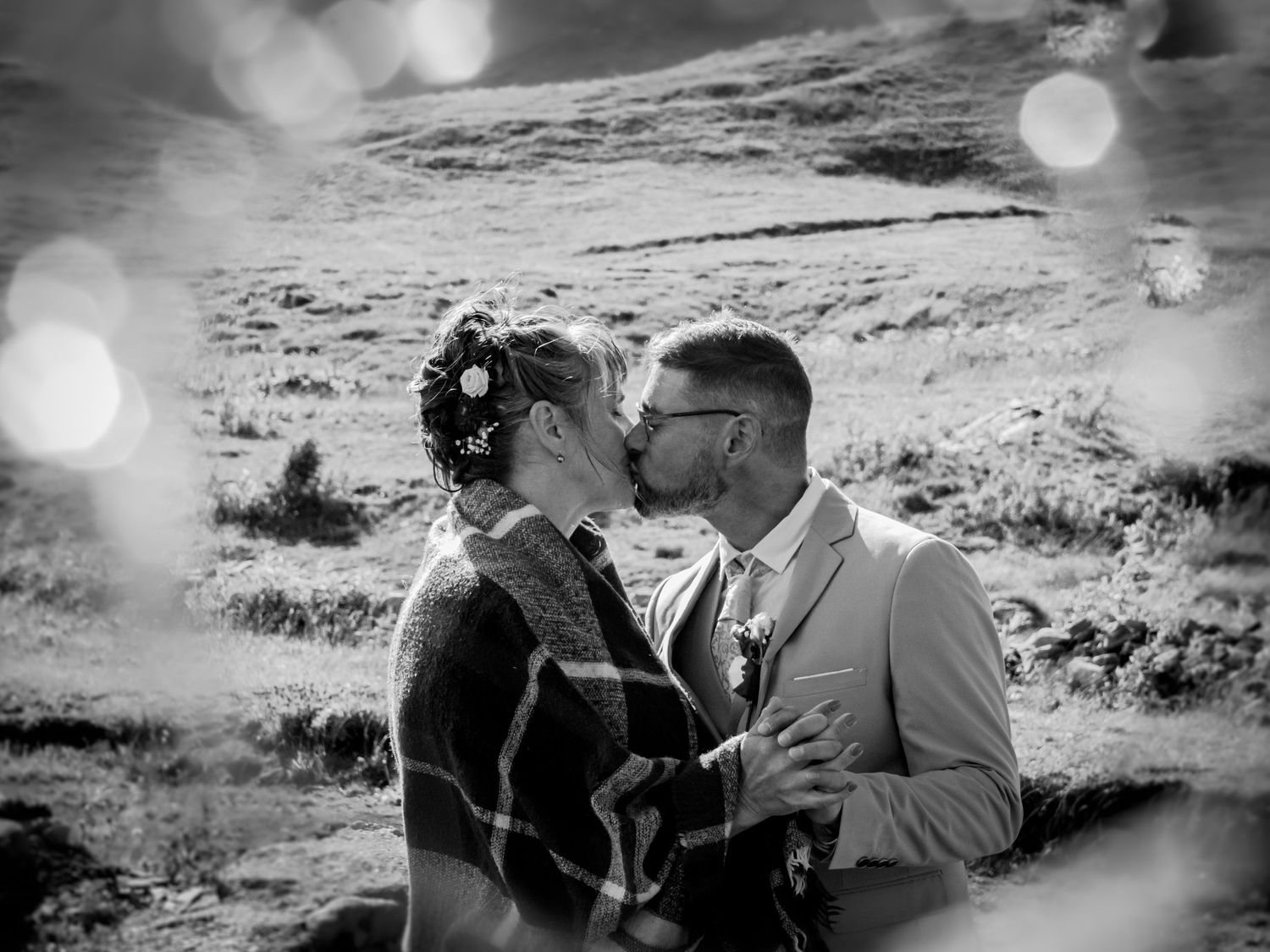 Photo de mariage en noir et blanc : couple s'embrassant en montagne, mariée avec plaid à carreaux et fleurs dans les cheveux, effet bokeh.