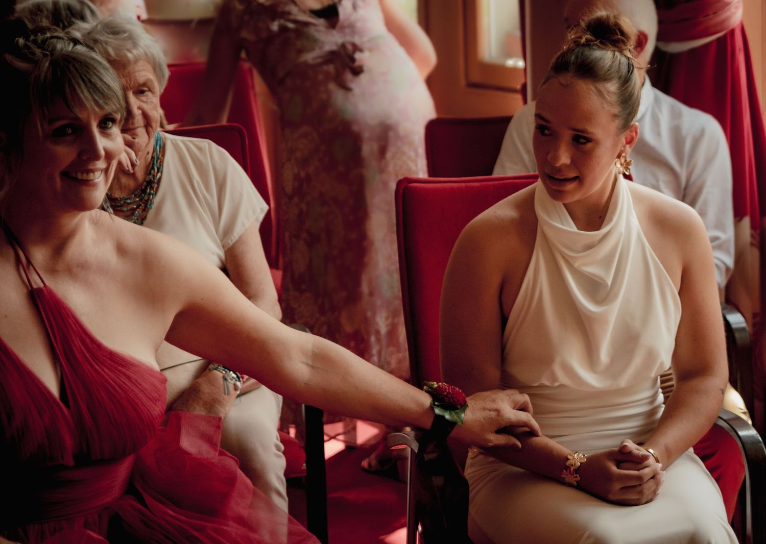 La mariée en robe rouge assise à côté de sa fille en robe blanche, lui tenant tendrement la main lors de la cérémonie de mariage, émotions !