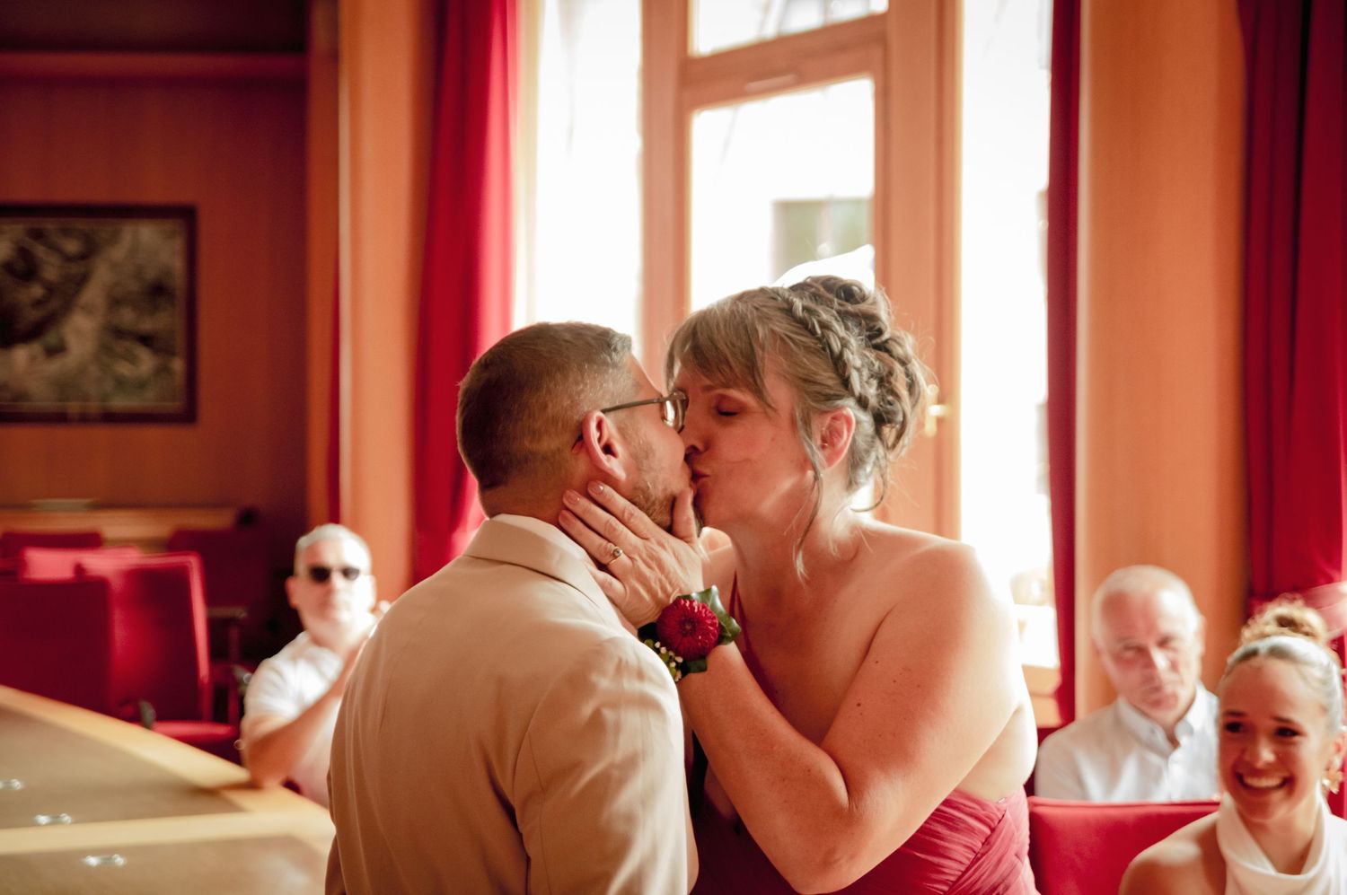 Baiser des mariés lors de la cérémonie civile en mairie, mariée en robe rouge et marié en costume beige, ambiance chaleureuse et émouvante.