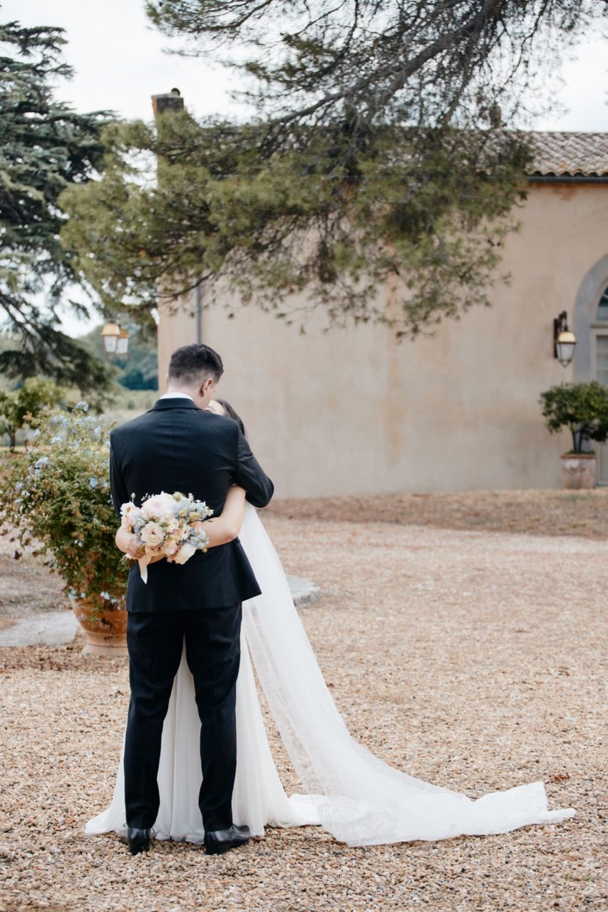A romantic wedding couple embraces outdoors at a rustic venue with a flowing white veil trailing behind them.