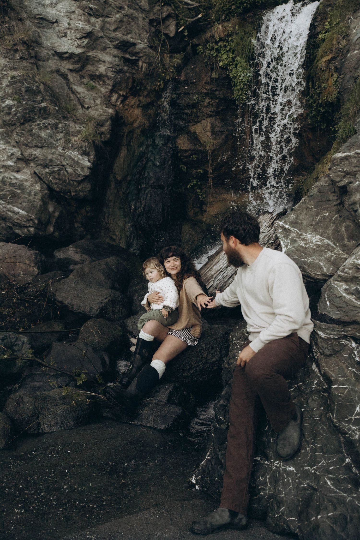 A wife reaching for her husband's hand while holding a baby near a waterfall during an intimate photoshoot