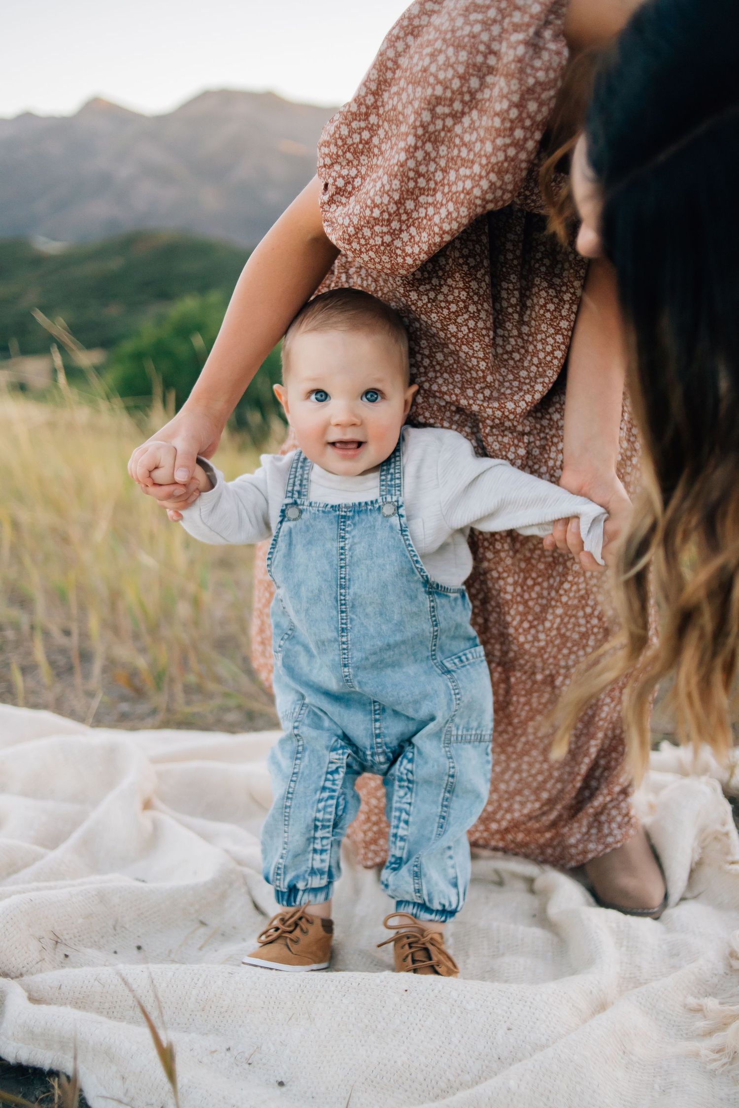 A baby learning to walk being helped by his mother during an outdoor family photo session at golden hour