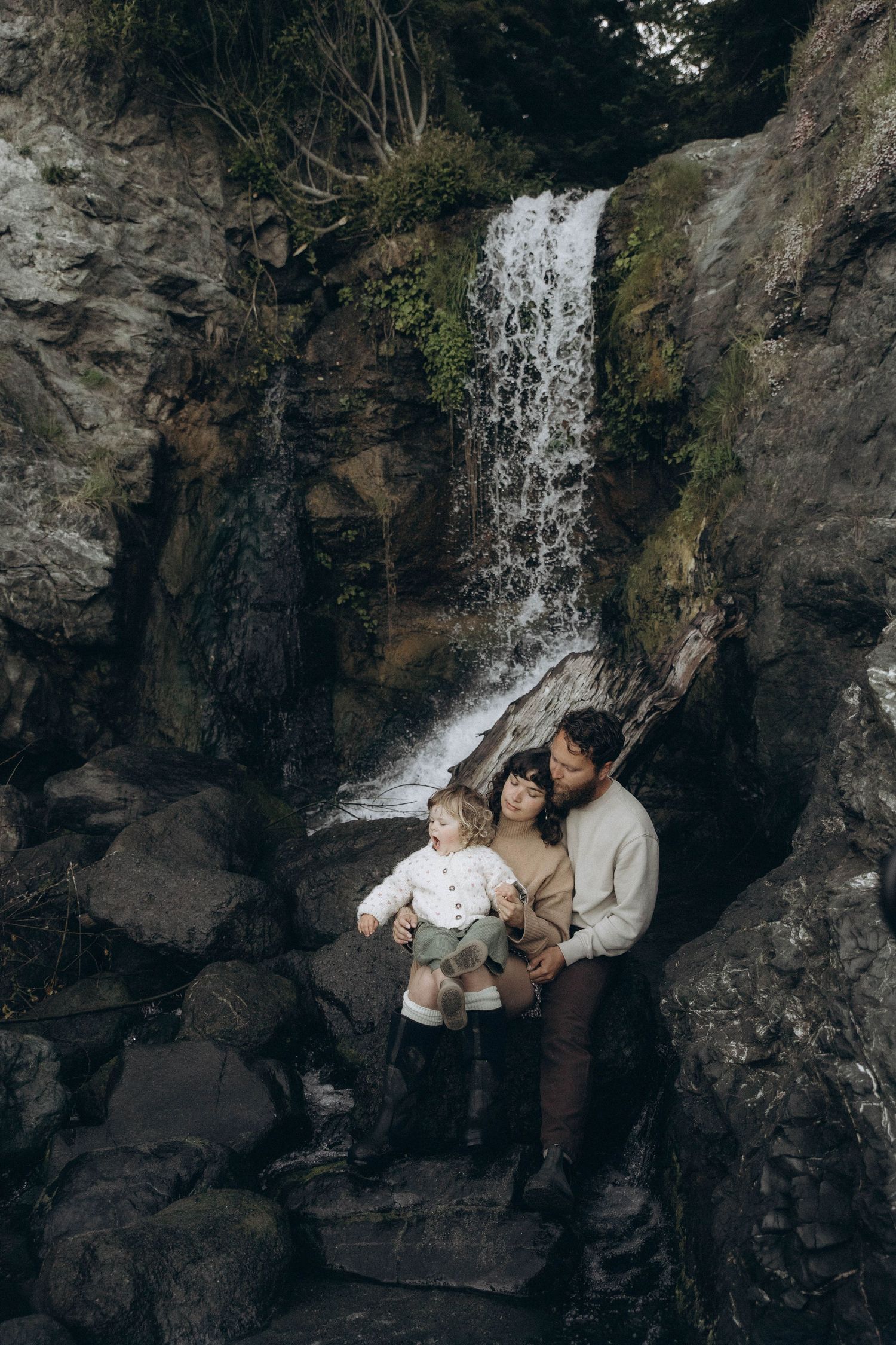 A mother and father cuddle their toddler while enjoying a waterfall during an early morning photoshoot