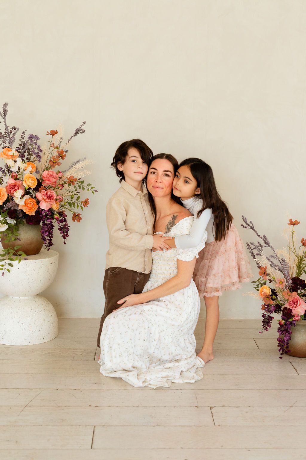A beautiful mother being embraced by her two toddlers in a white studio surrounded by flowers during a mommy and me mini photoshoot