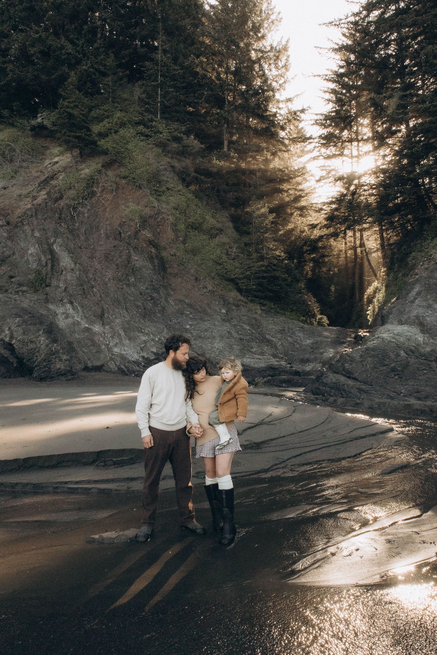 A family of three enjoy a sunrise photo session on the beach during sunrise as the light gently filters through the trees