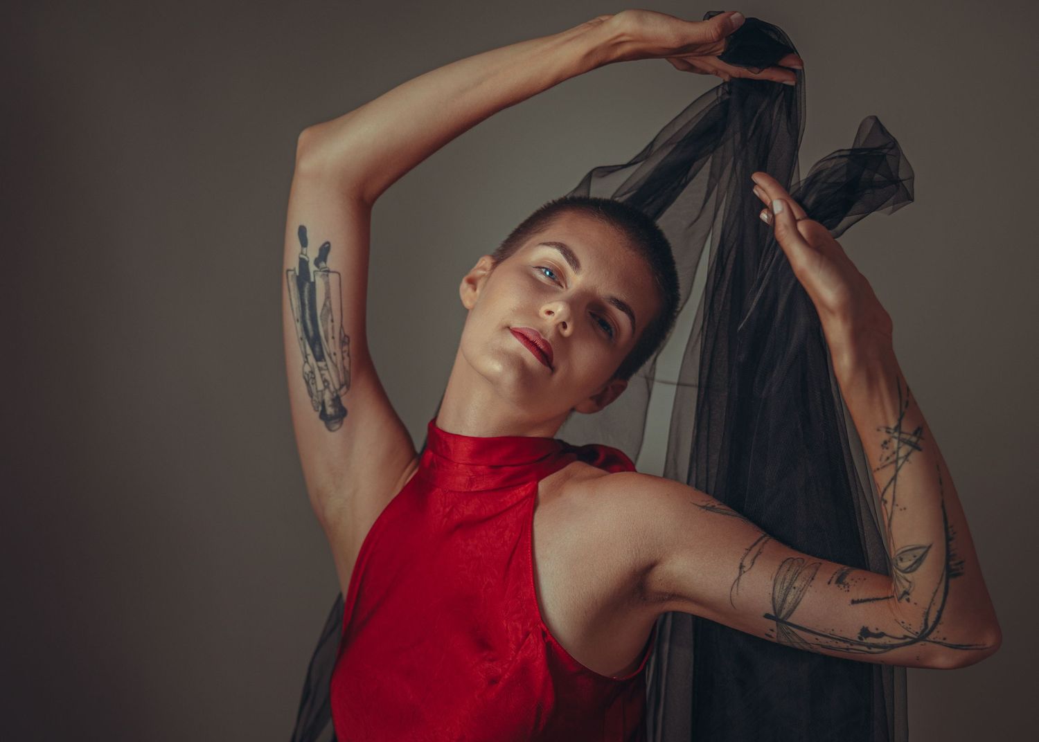 Person in red top with tattoos holding black veil overhead against gray background.