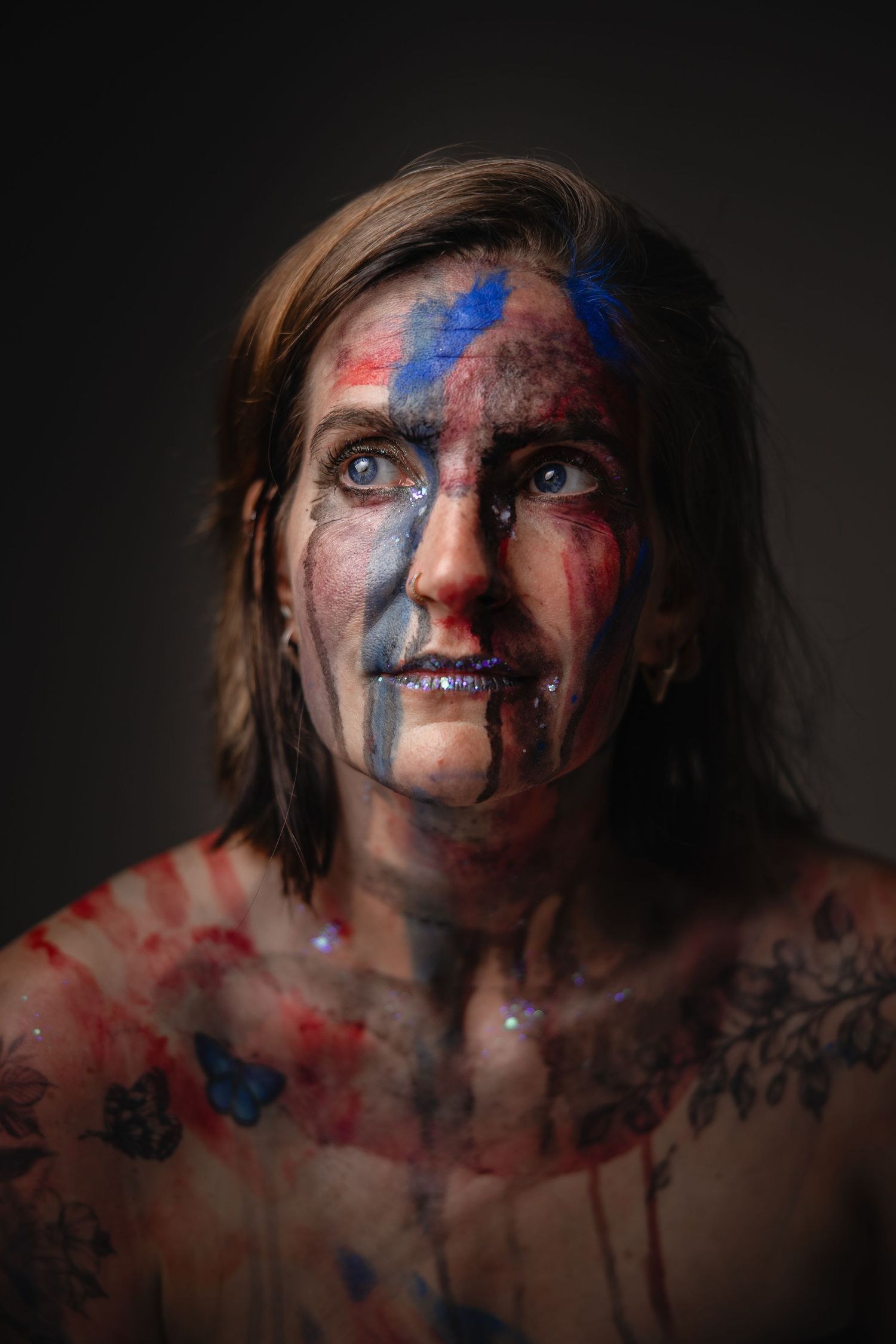 Portrait of a person with blue and red face paint, tattoos, and glitter, against a dark background.