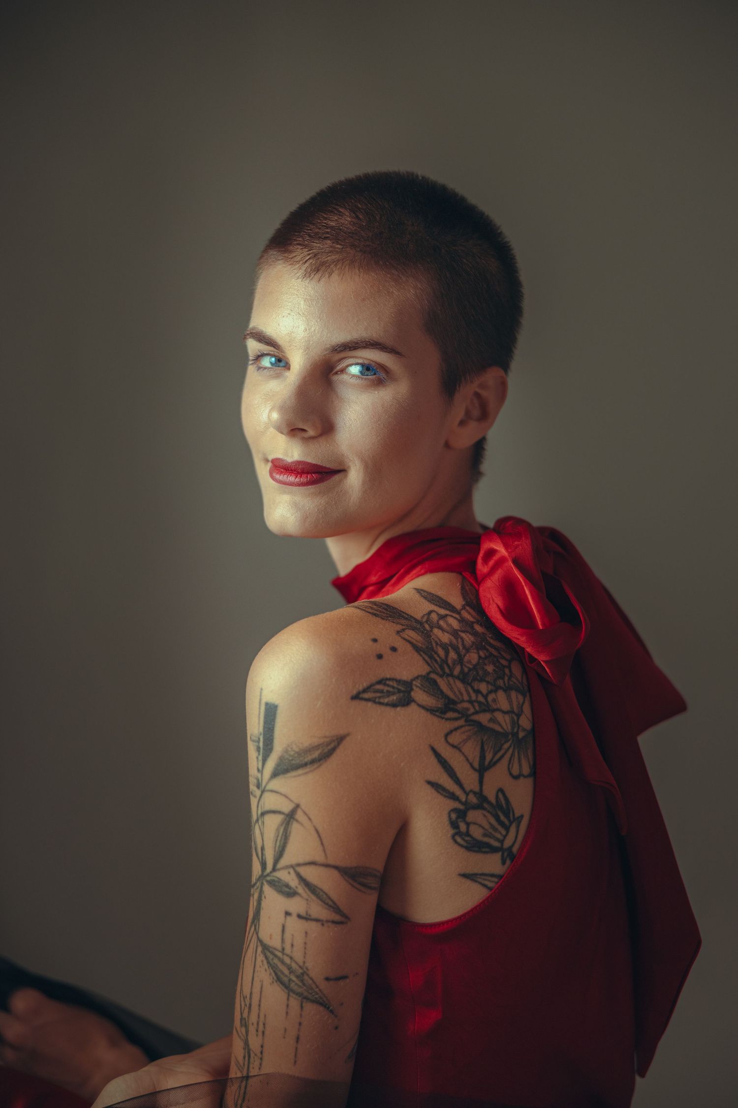 Woman with shaved head, blue eyes, red lips, and floral arm tattoos wearing a red backless dress with bow.
