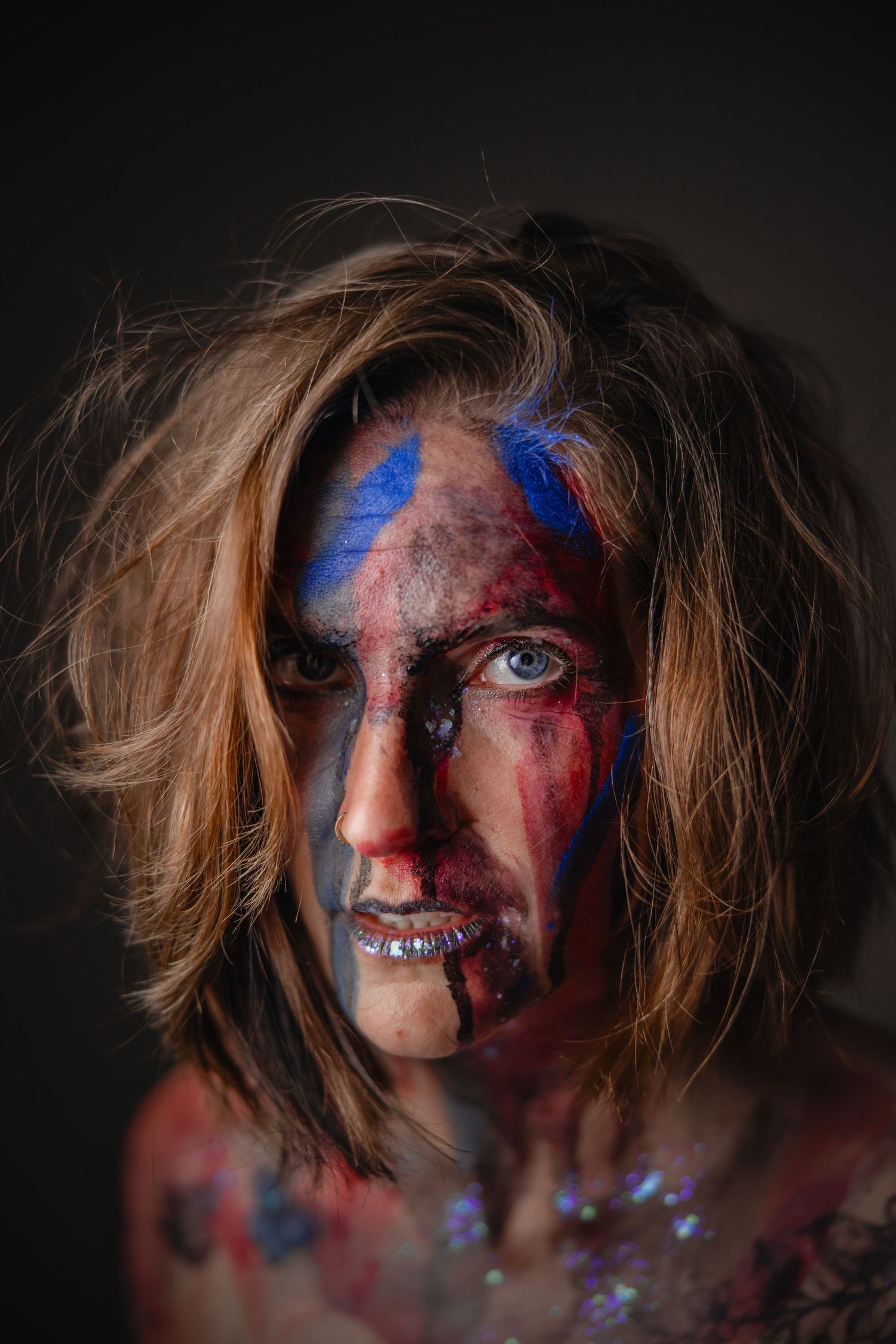 Person with dramatic Halloween makeup featuring blue and red face paint, messy hair, and glitter on dark background.