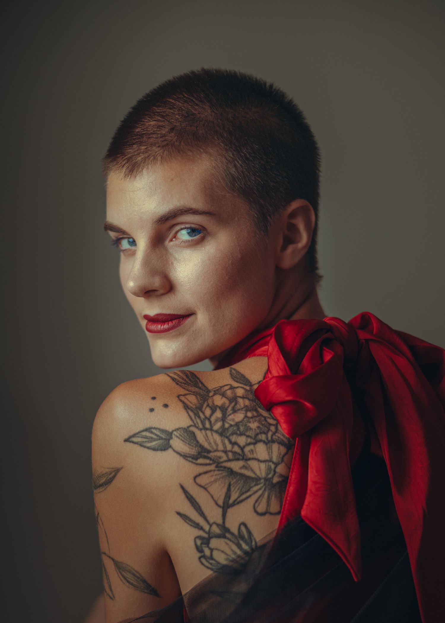 Portrait of a shaved-head woman with tattoos, red lips, and a red silk bow draped over her shoulder.