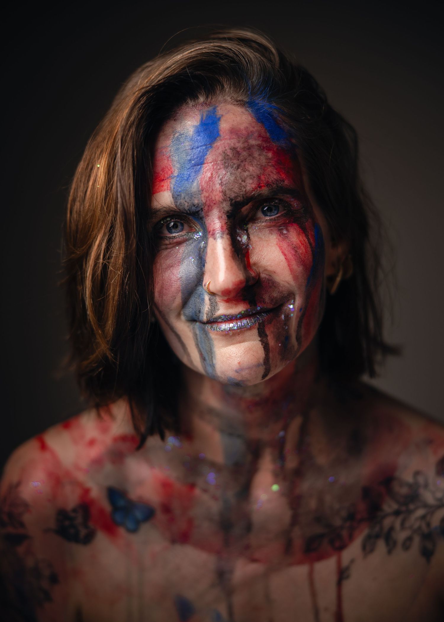 Person with colorful blue and red face paint smiling, tattoos visible on chest, artistic portrait photography.