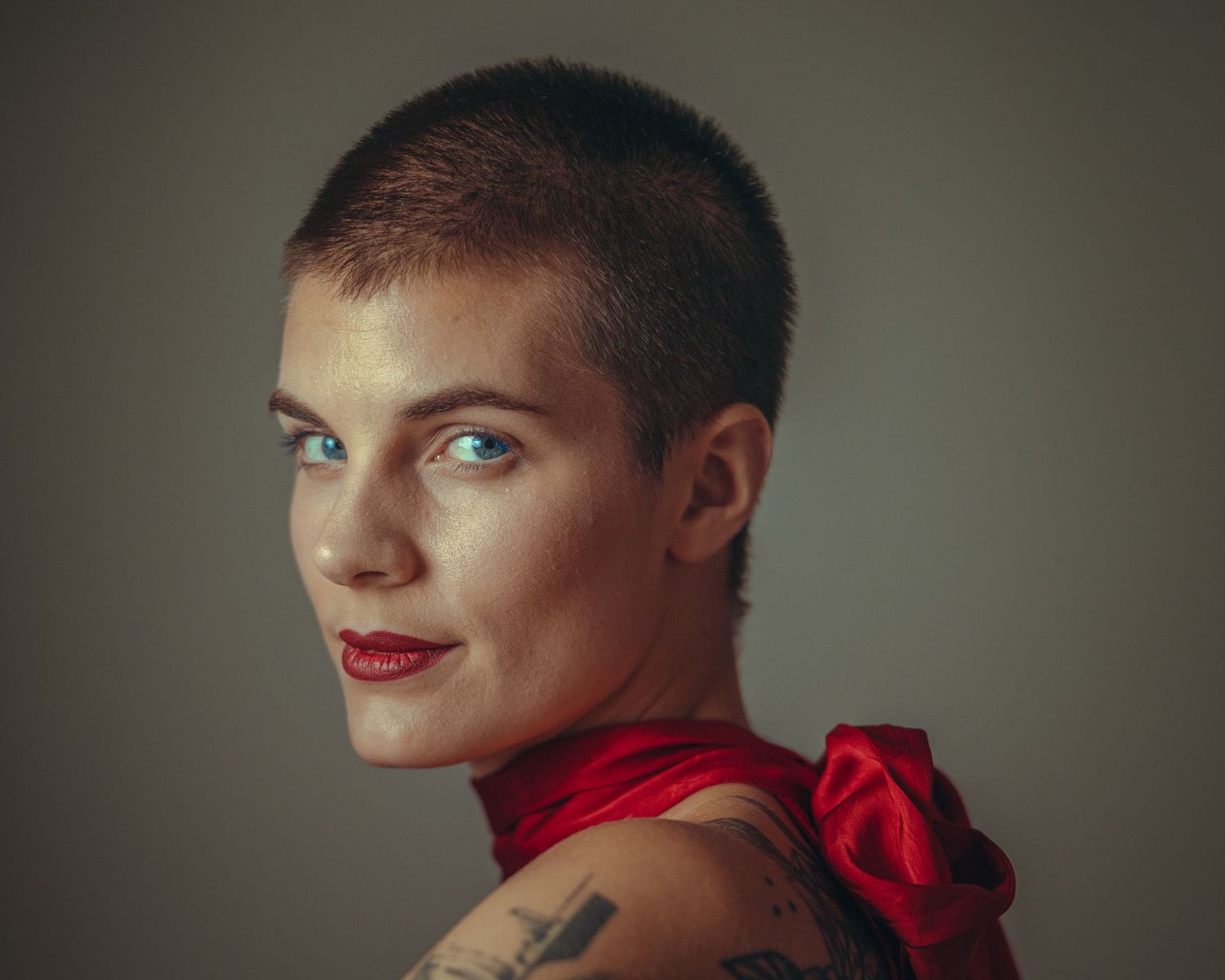Woman with buzz cut, blue eyes, red lips, and shoulder tattoo wearing a red top, portrait photo.