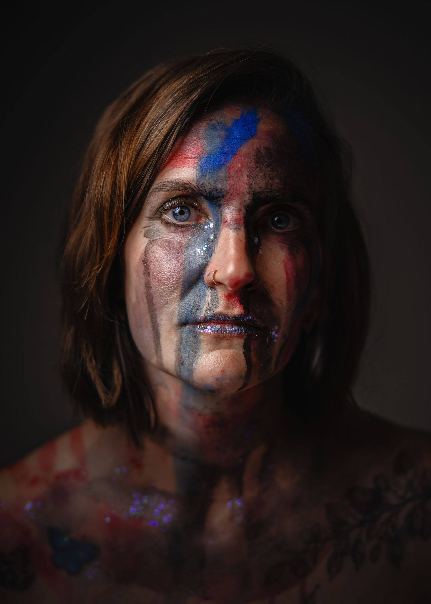 Person with colorful blue and red paint on face and body, dramatic studio portrait with dark background.