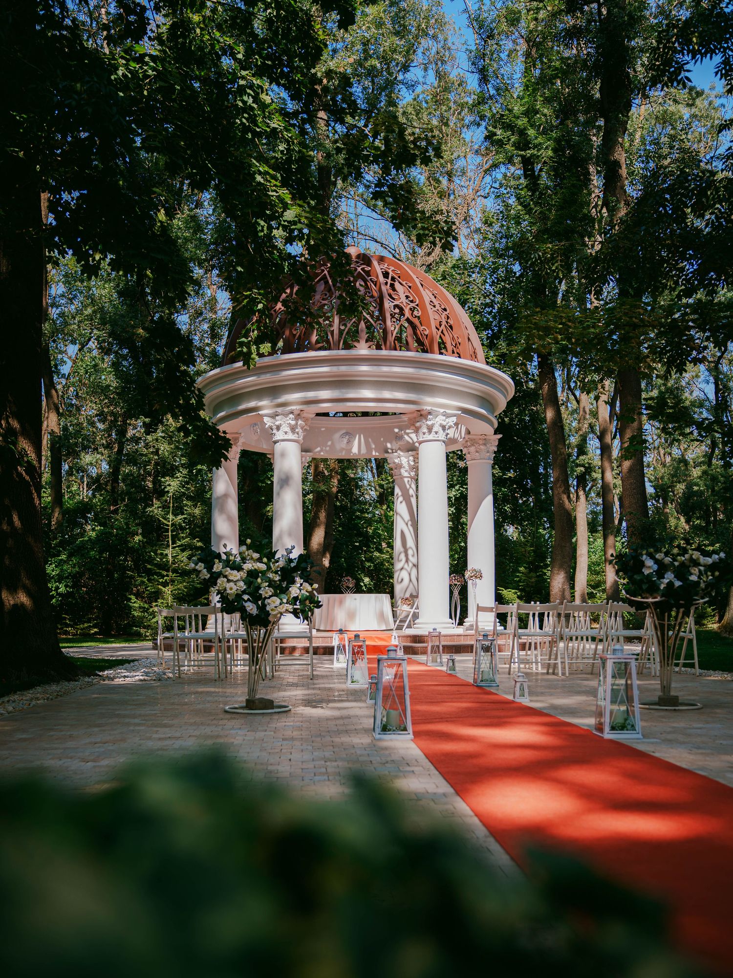 Elegant outdoor wedding ceremony with red carpet aisle leading to white columned gazebo with ornate dome in lush garden.