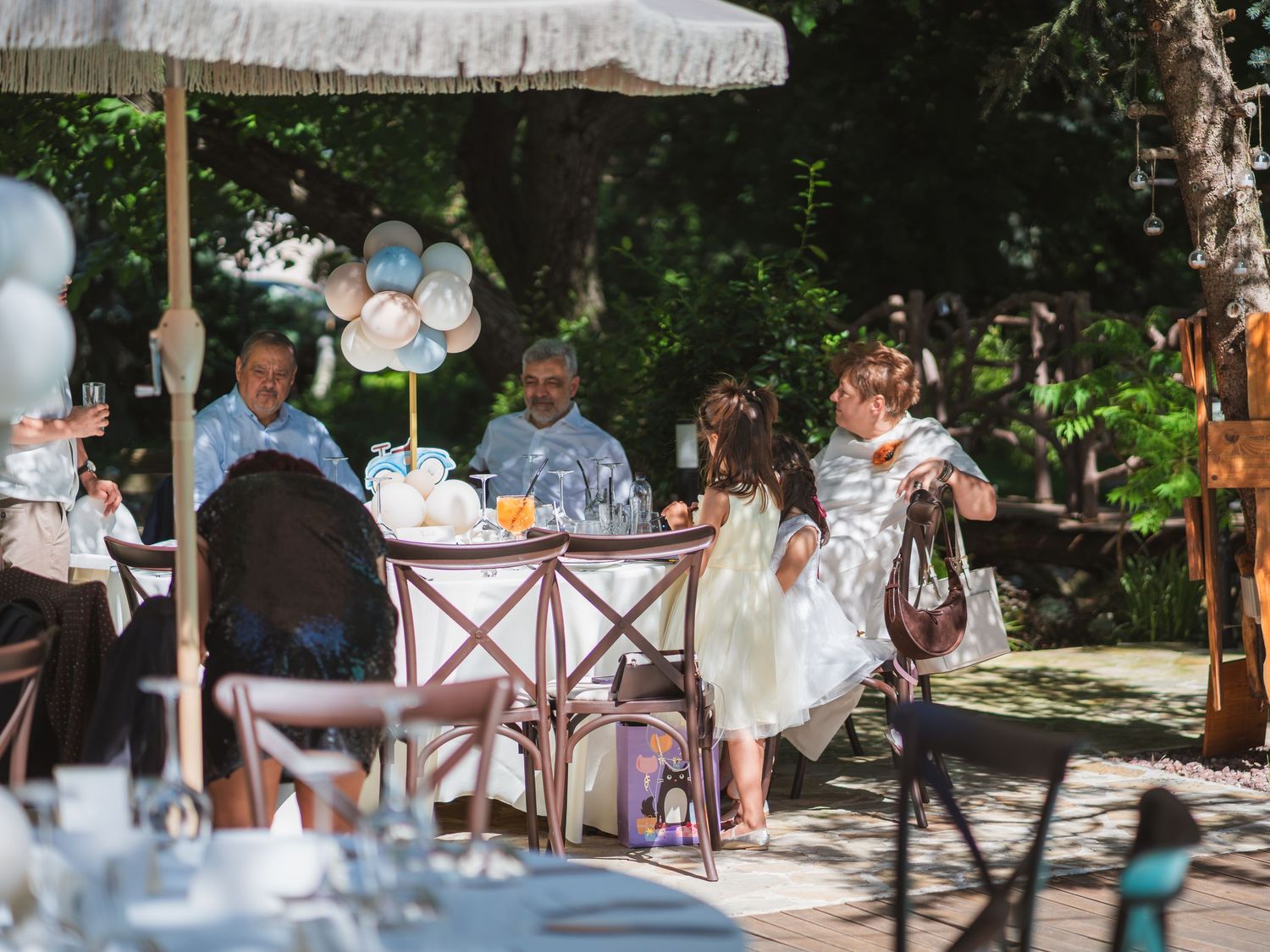 Invitati asezati la mesele de botez in aer liber decorate cu baloane albastre si albe
