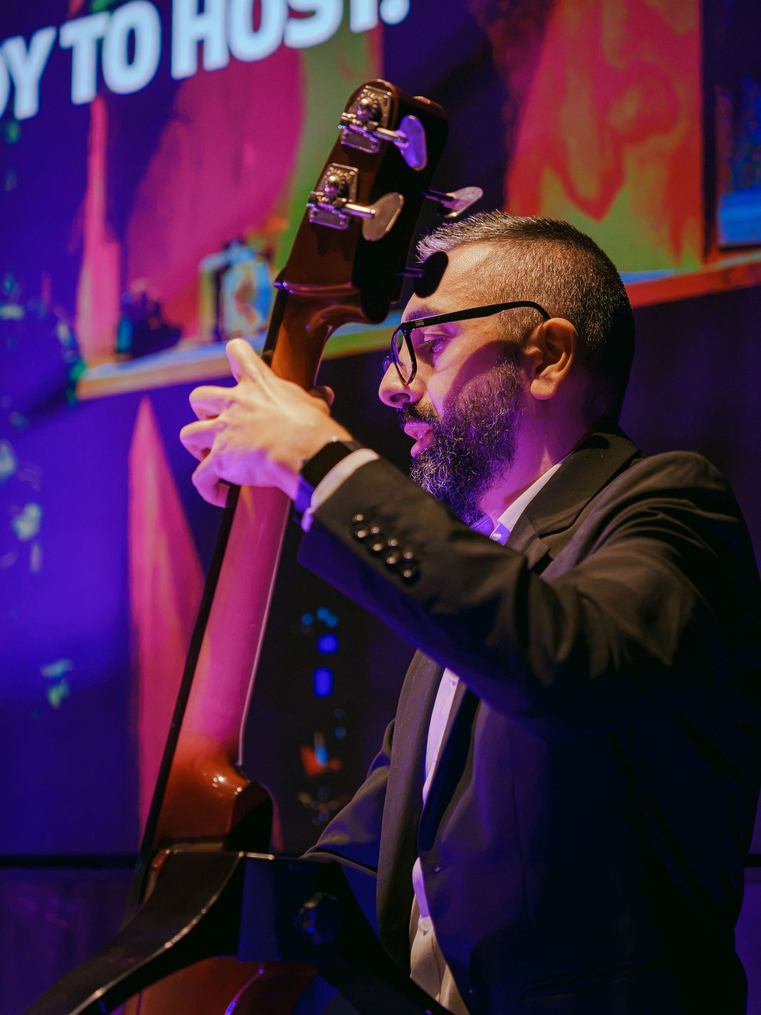 A bearded man in a black suit plays double bass under colorful stage lighting at a live music event.