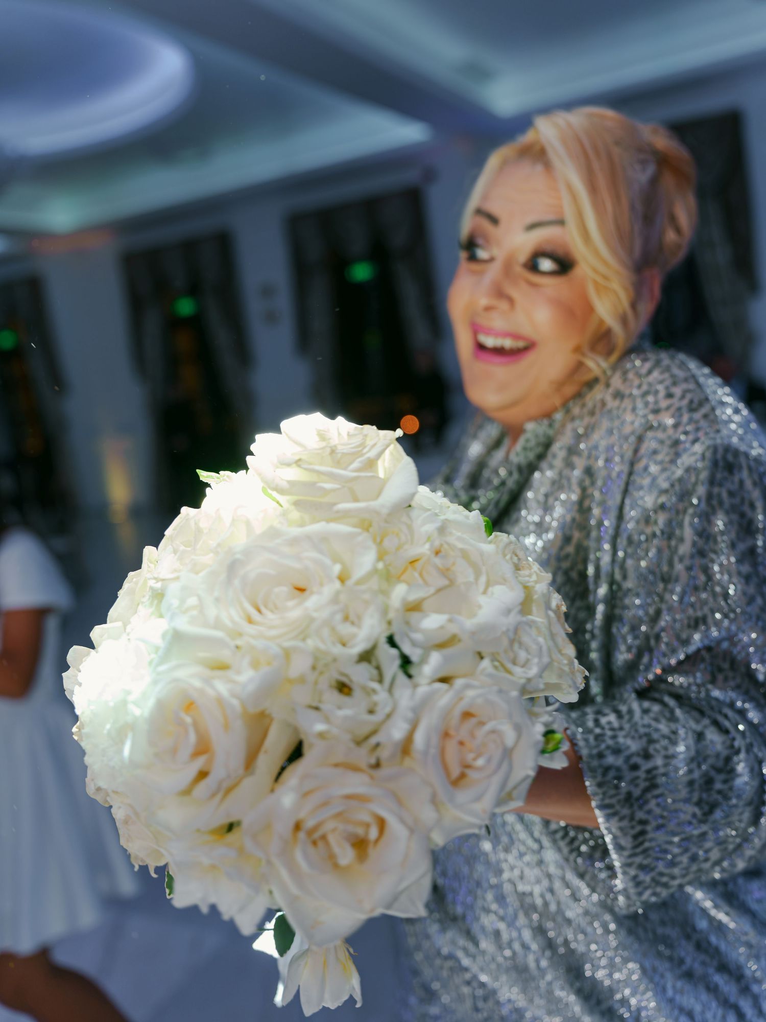 Smiling blonde woman in silver sequin dress holding elegant white rose bouquet at wedding reception.