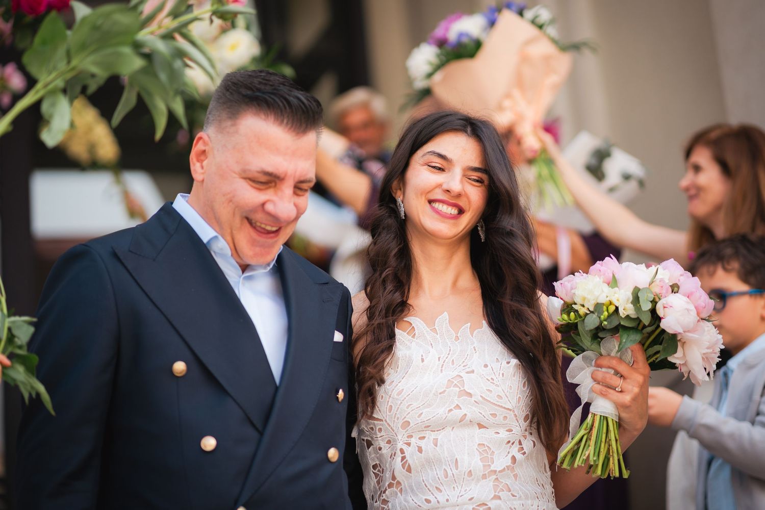Smiling bride holding a bouquet stands beside groom in navy blazer outside wedding venue.